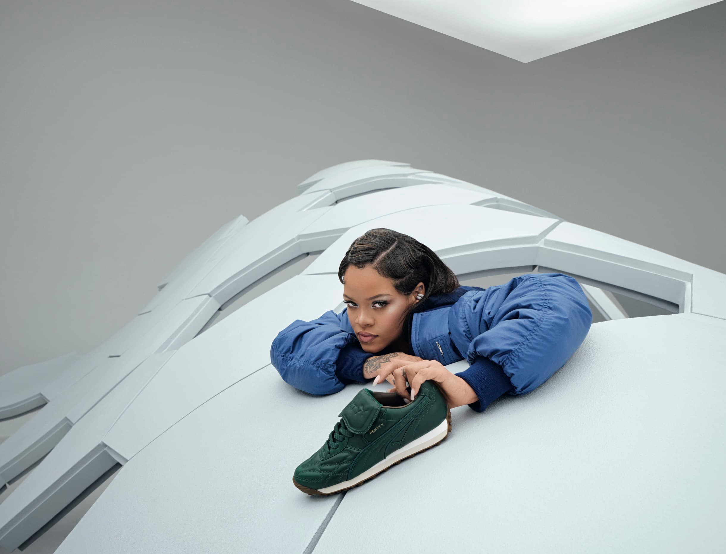 Rihanna lies on a geometric white structure in a blue jacket, presenting a green FENTY x PUMA sneaker to the camera.
