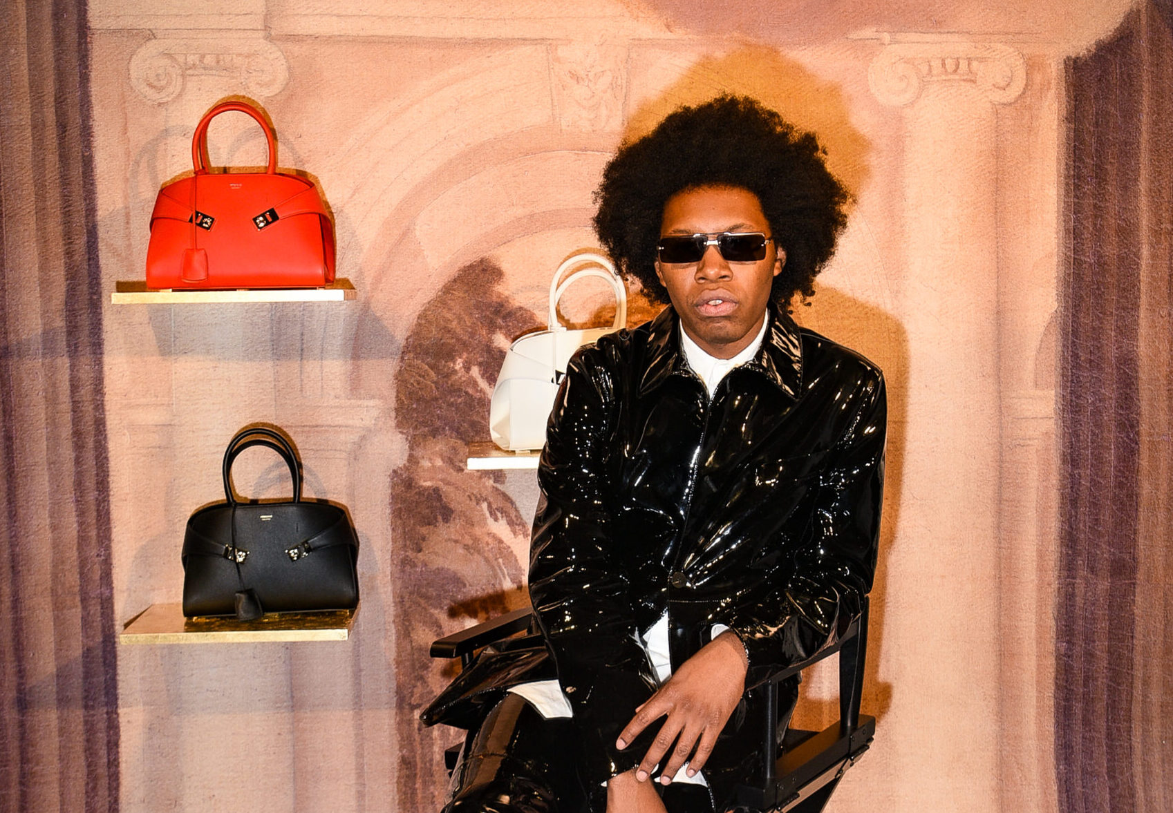 Jeremy O'Harris exudes style at the Ferragamo Fall-Winter 2023 event, seated amidst the latest handbags, wearing a glossy black ensemble and sunglasses.