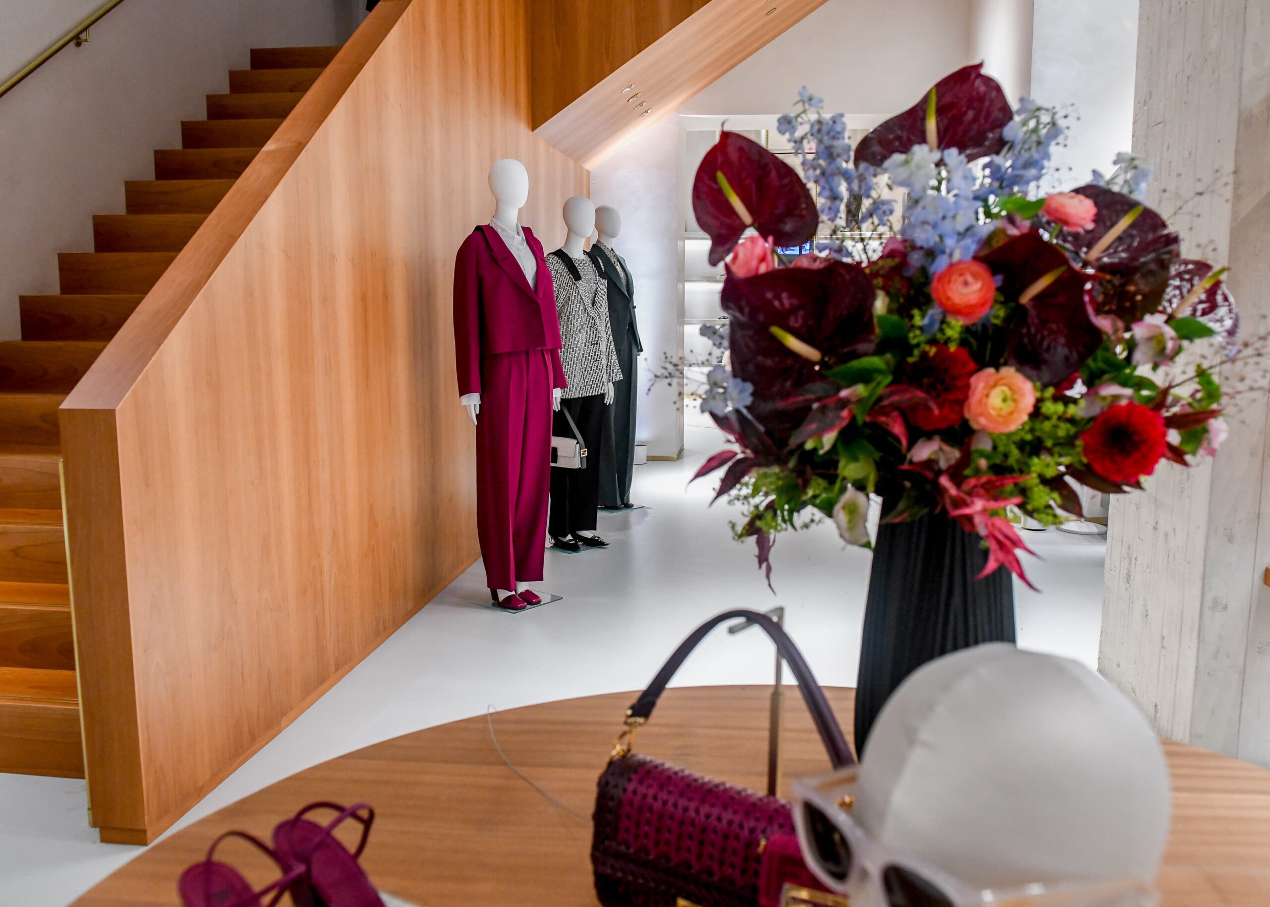 Inside a modern boutique, mannequins display designer outfits next to a wooden staircase, with a foreground of vibrant floral arrangements and luxury handbags.
