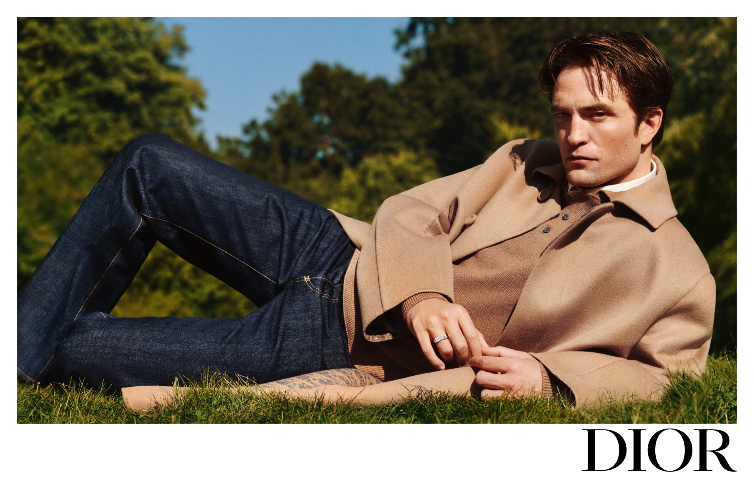 Robert Pattinson lounges on the grass in a DIOR campaign, wearing a tan jacket and blue jeans, conveying a relaxed yet sophisticated style.