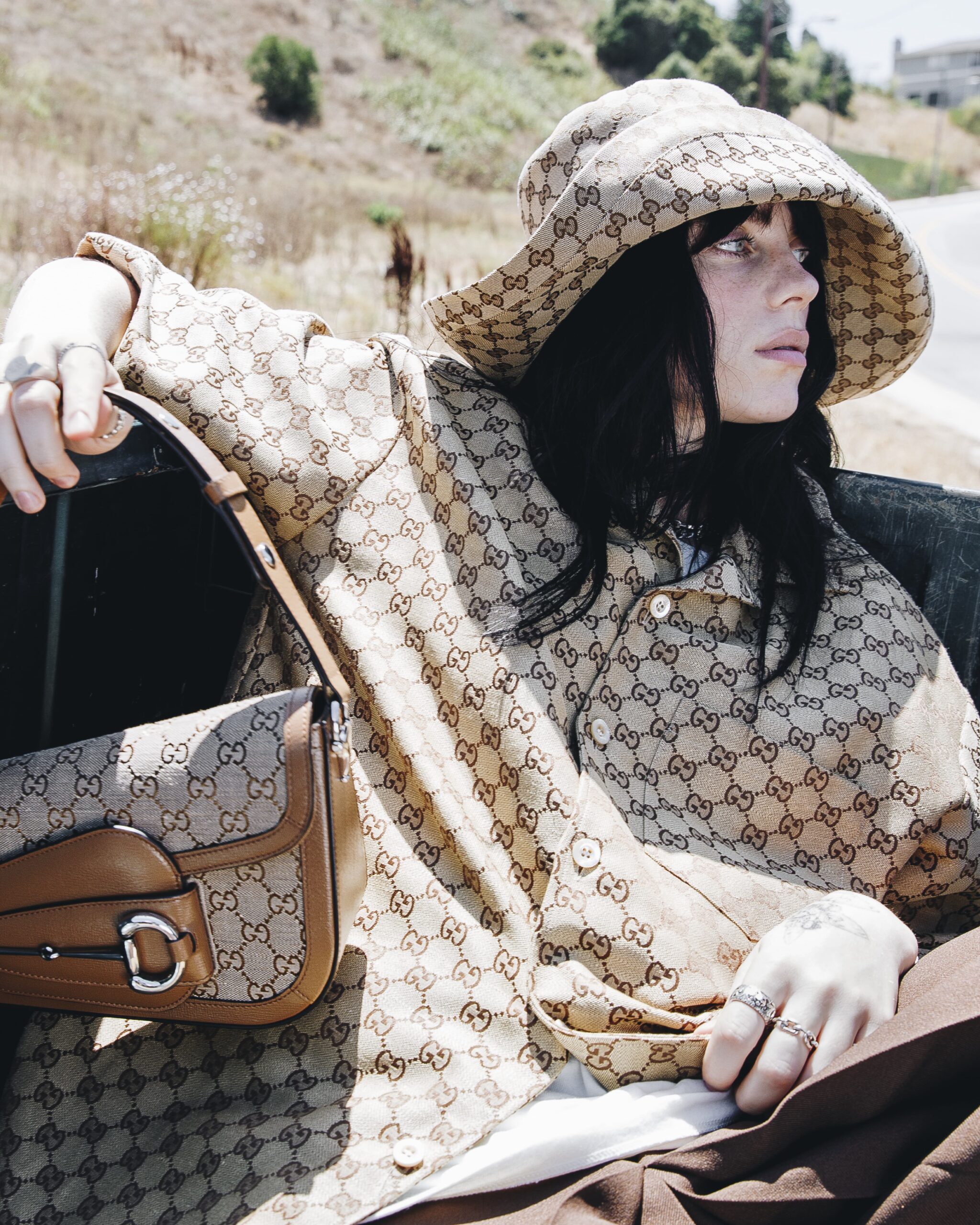 Billie Eilish is featured in a Gucci campaign, donning a monogrammed bucket hat and matching coat with the Horsebit 1955 bag, set against a natural backdrop.