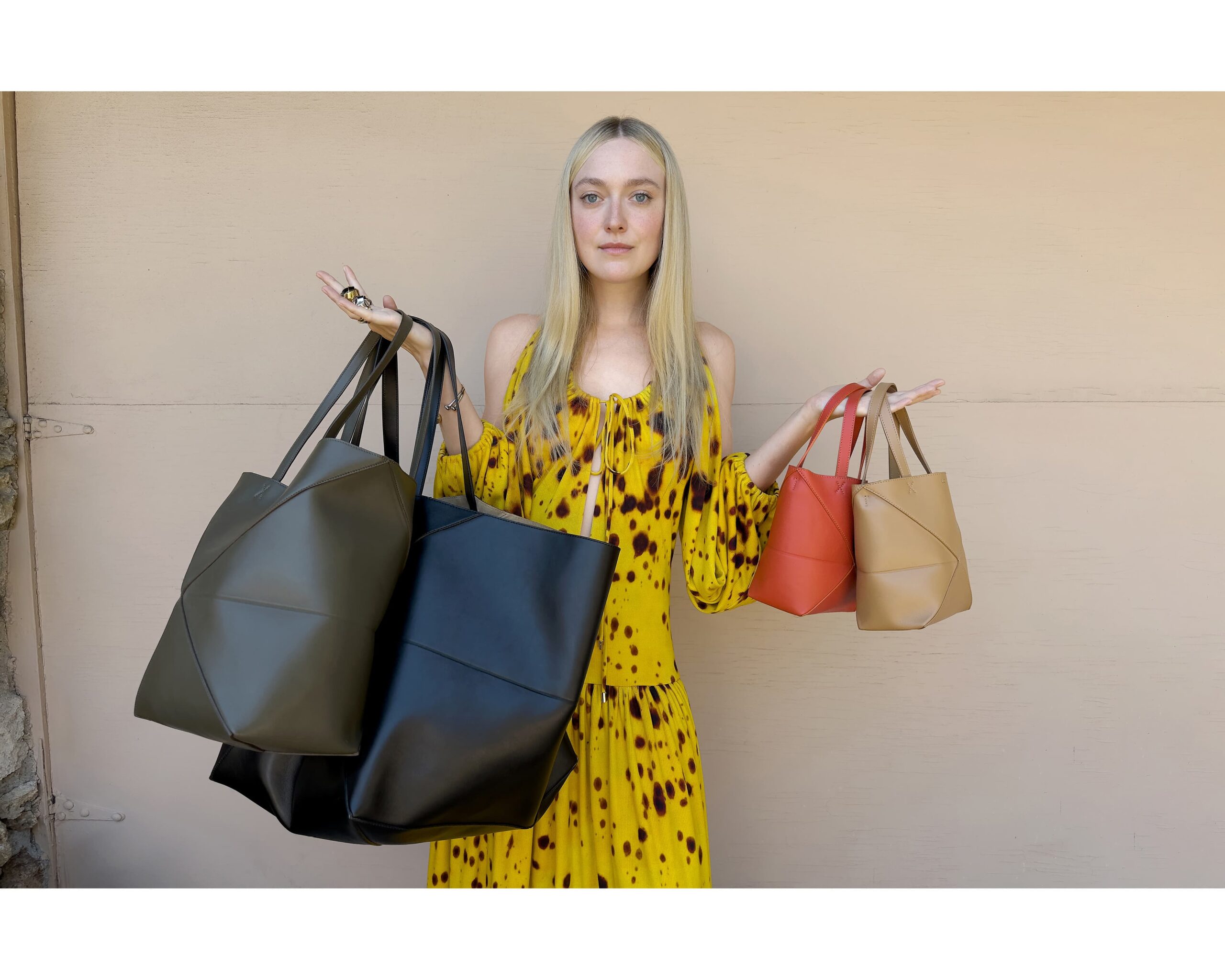 Dakota Fanning in a yellow patterned dress stands against a plain wall, holding three LOEWE tote bags in varying sizes and colors.