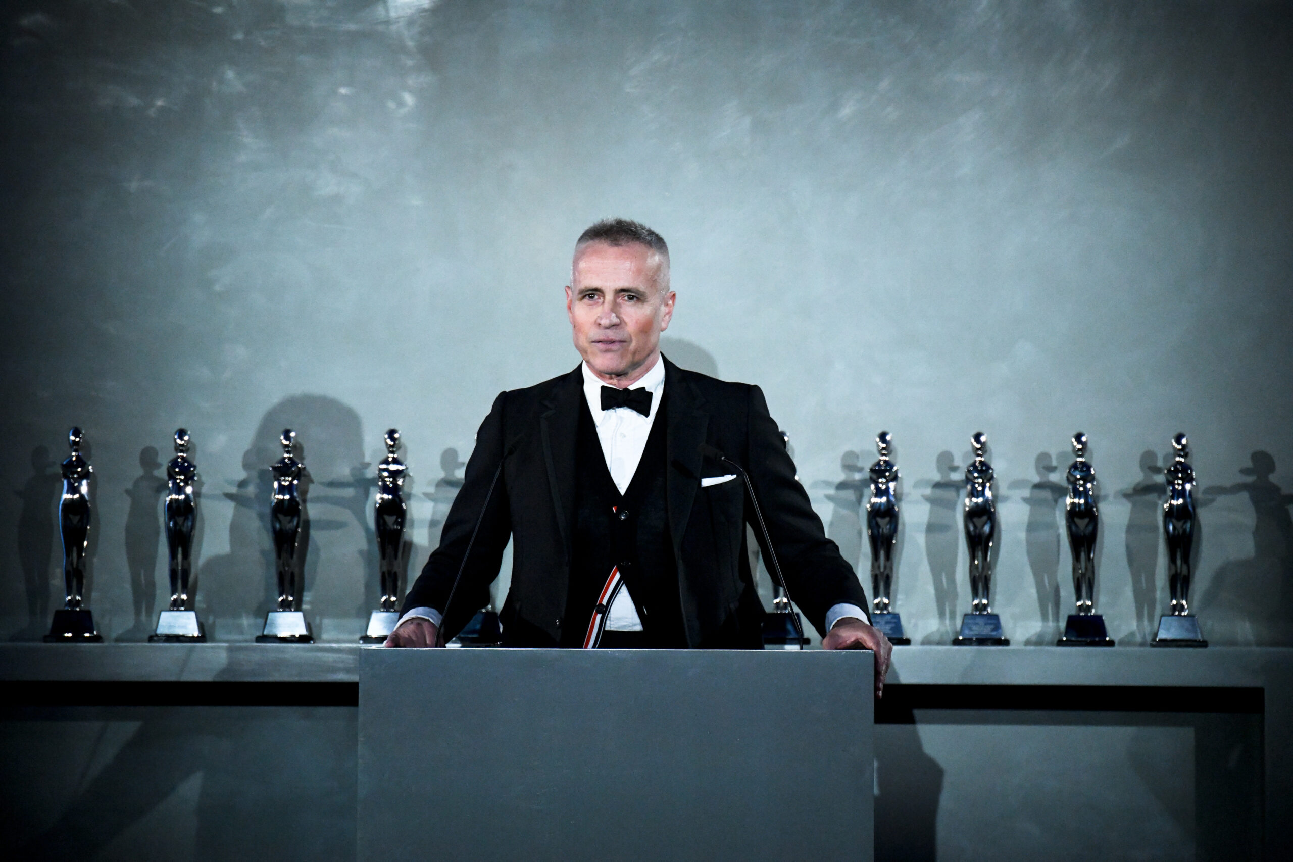 CFDA Chairman Thom Browne stands ready at the podium, with the evening's coveted awards lined up behind him, set to celebrate the industry's top talents at the prestigious CFDA Fashion Awards presented by Amazon.