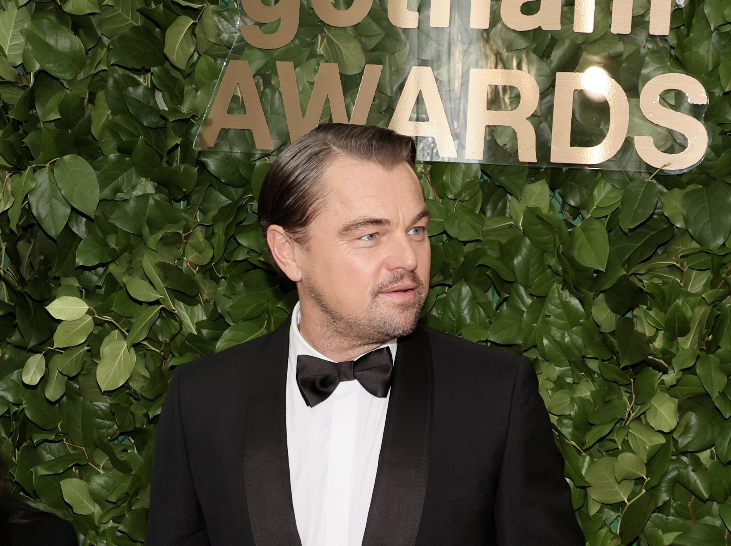 Leonardo DiCaprio attends the 33rd Annual Gotham Awards, dressed in a classic black tuxedo with a bow tie, against a backdrop of lush green foliage.