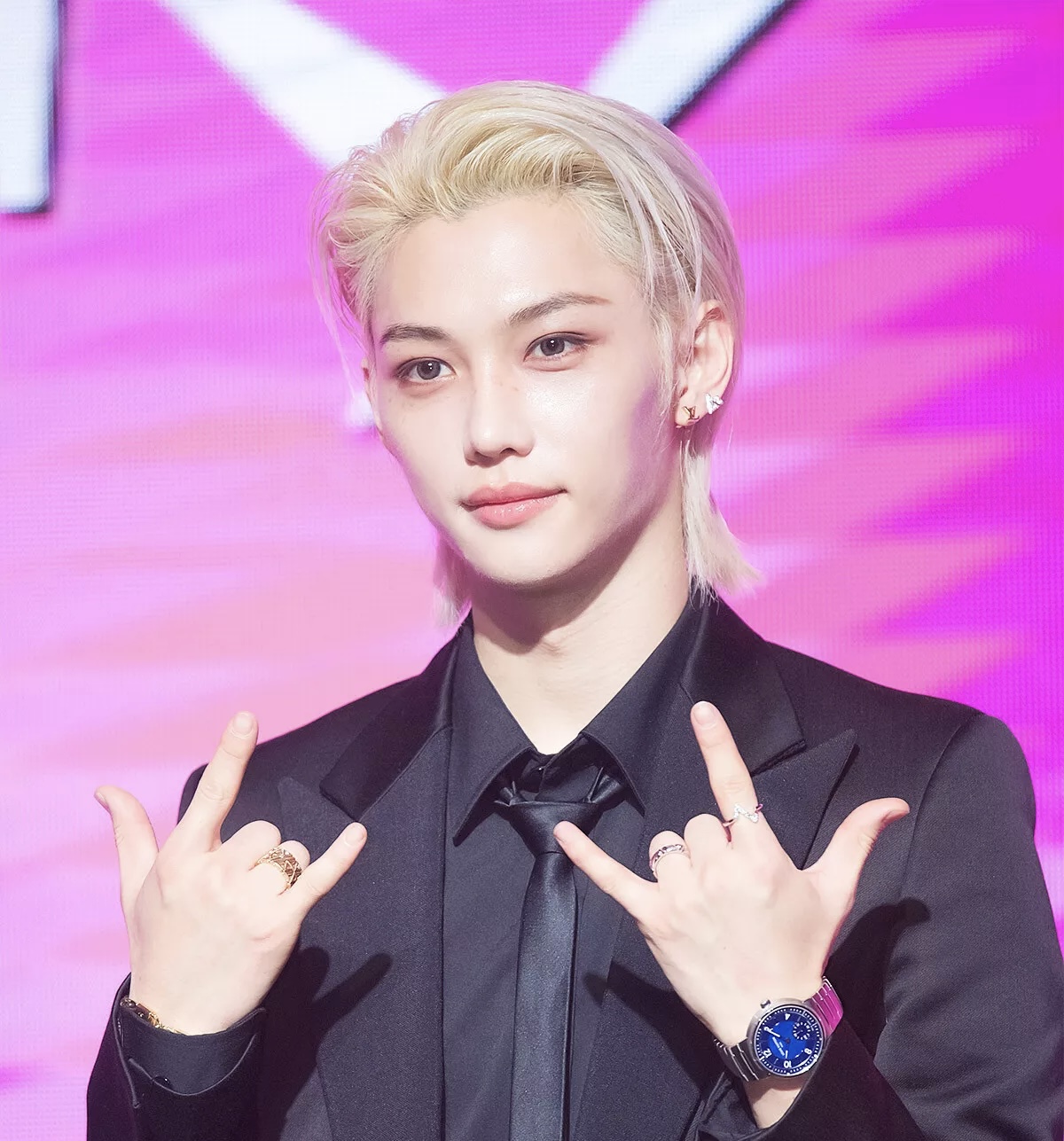 Felix of Stray Kids at a press conference, dressed in a Louis Vuitton black tuxedo and accessorized with fine jewelry, against a pink backdrop.