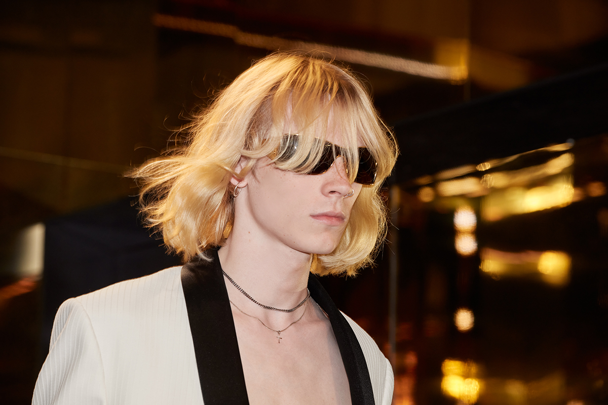 A close-up of a model from the Celine Homme Summer 24 collection, featuring a blond shaggy hairstyle, modern sunglasses, and a white jacket with contrasting lapels. Details include a slim black tie and a delicate cross pendant necklace against a golden mirrored backdrop.