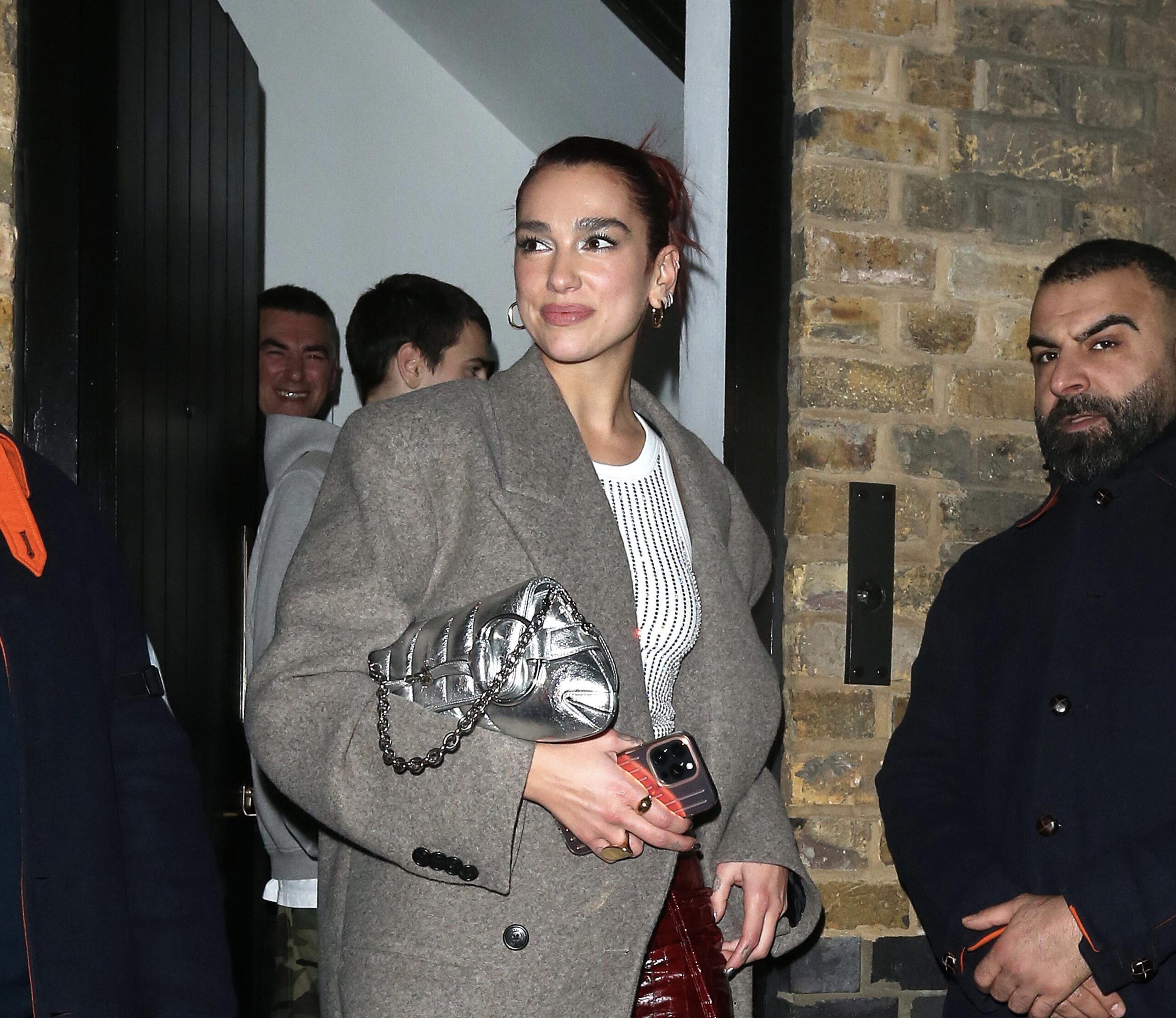 Dua Lipa, leaving The Chiltern Firehouse in London, combines classic and contemporary elements with a grey overcoat, metallic clutch, red vinyl trousers, and white-heeled boots, creating a look that is both timeless and modern.