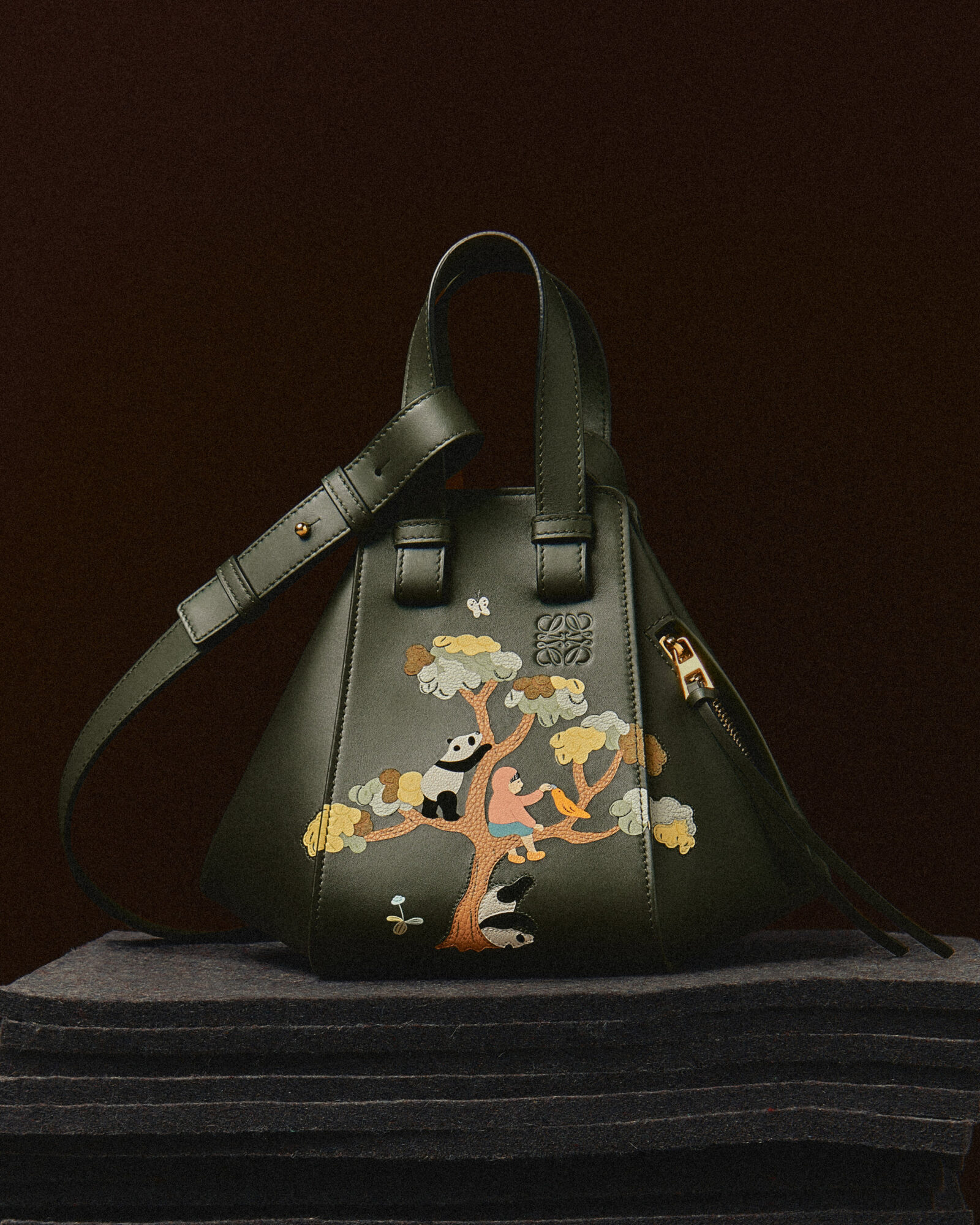 Introducing the LOEWE Hammock Bag: A Festive Ode to Craftsmanship - This deep-green leather masterpiece showcases a whimsical scene of playful pandas amid a forest, intricately brought to life with leather marquetry and hand-painting techniques, exemplifying the brand's commitment to storytelling through fashion.