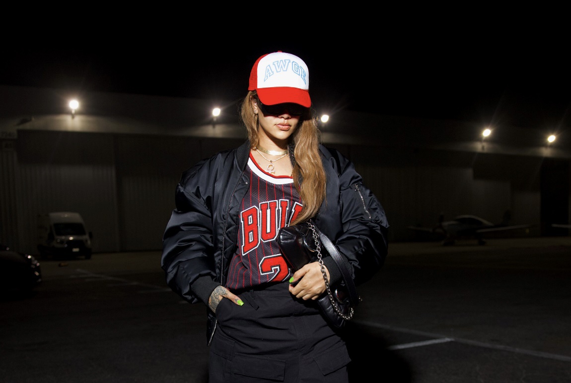Rihanna, in a red "AWGE" cap and a "BULLS 2" jersey, exudes a sporty yet glamorous vibe. She accessorizes with a black Gucci Horsebit Chain bag and finishes the look with an oversized bomber jacket and split skirt, set against a nocturnal Las Vegas parking lot backdrop.