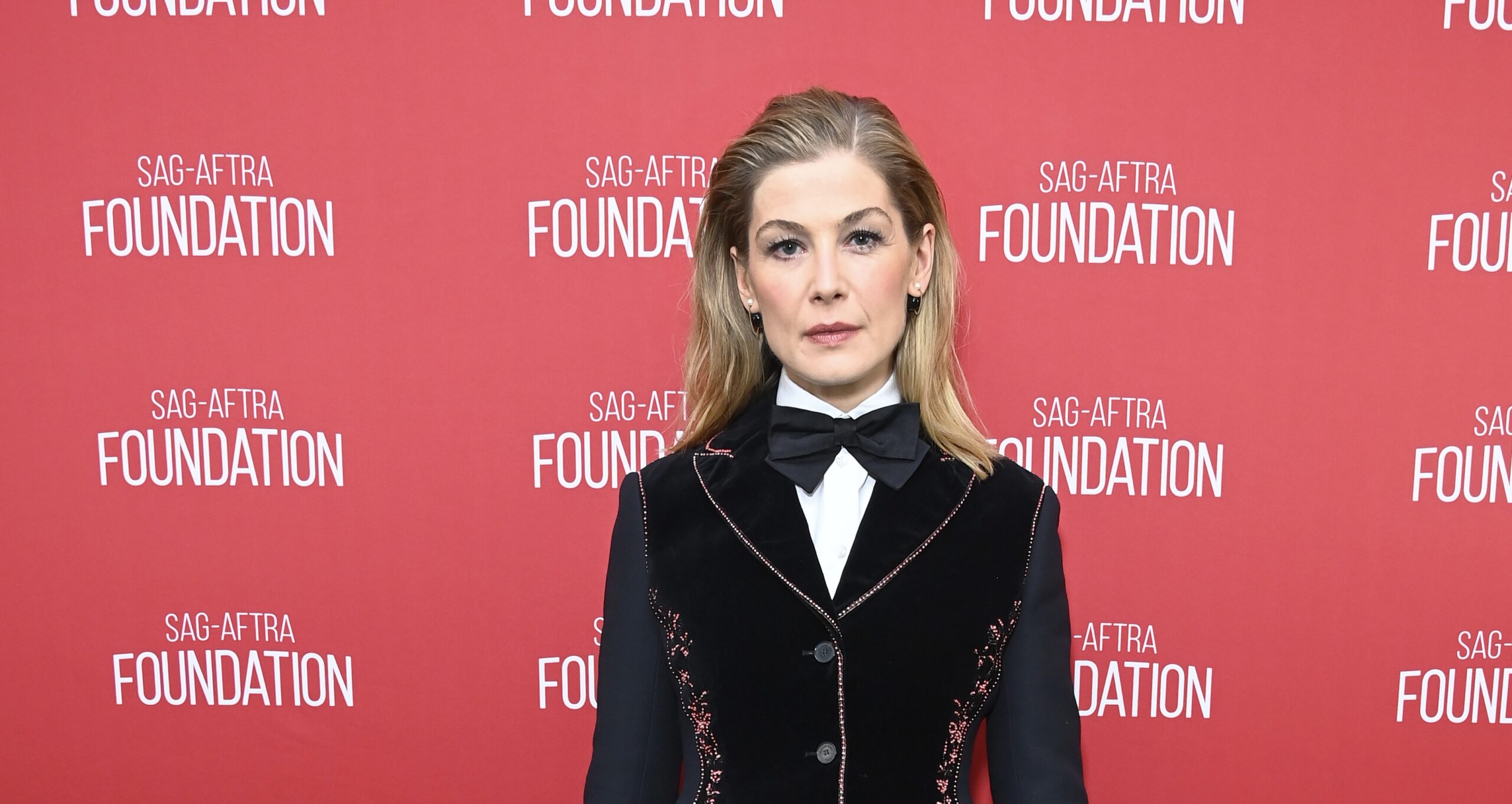 Actress Rosamund Pike at the SAG-AFTRA Foundation event in Los Angeles, wearing a black velvet dress with pink embroidery, a white high-collar shirt, and a black bow tie.