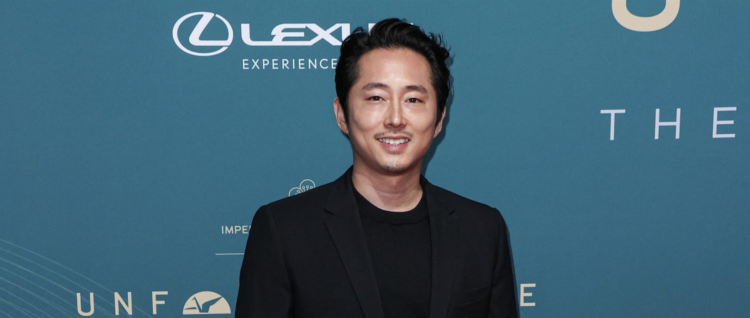 Actor Steven Yeun at the Unforgettable Gala Asian American Awards in Beverly Hills, dressed in an elegant all-black suit with a satin lapel, smiling on the red carpet.