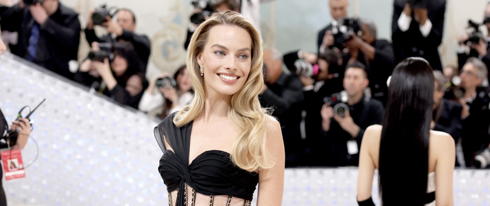 Margot Robbie exudes elegance in a black cut-out gown with golden accents, smiling warmly at a prestigious gala event. Her blonde hair is styled in gentle waves, and her poised demeanor is the epitome of refined style.