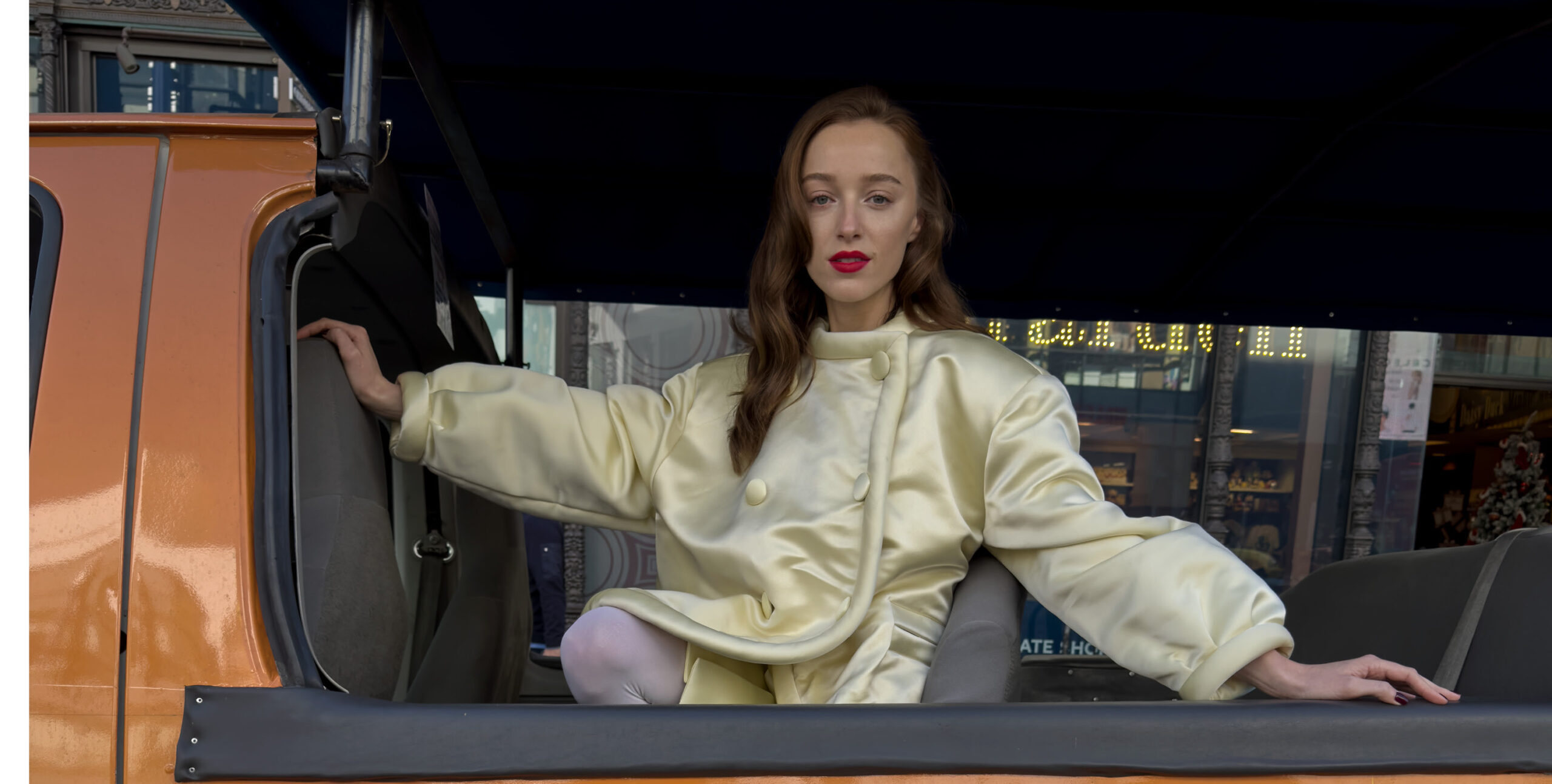 Phoebe Dynevor strikes a pose for W Magazine, sitting elegantly in an open vehicle. She wears a pale yellow satin jacket with bold button details, adding a touch of soft glamour to the urban setting. Her red lipstick contrasts with her light outfit, and her relaxed yet poised demeanor exudes a modern sophistication. Behind her, the city life blurs into the background, focusing all attention on her striking figure.