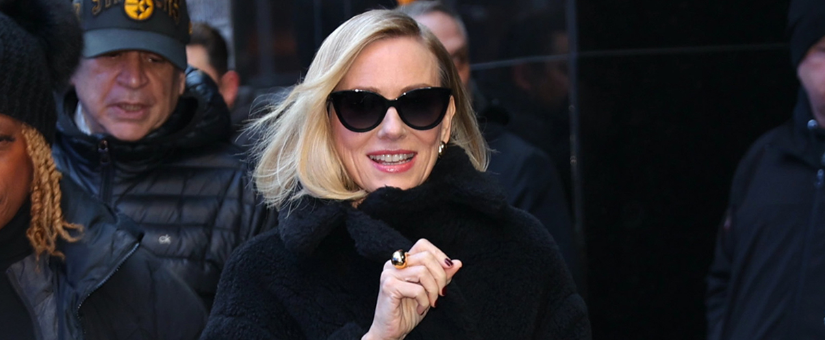Naomi Watts smiles while exiting a building in New York City, wearing a chic black fur coat, cream-colored skirt, and dark sunglasses, carrying a sleek CELINE Classique 16 Bag.