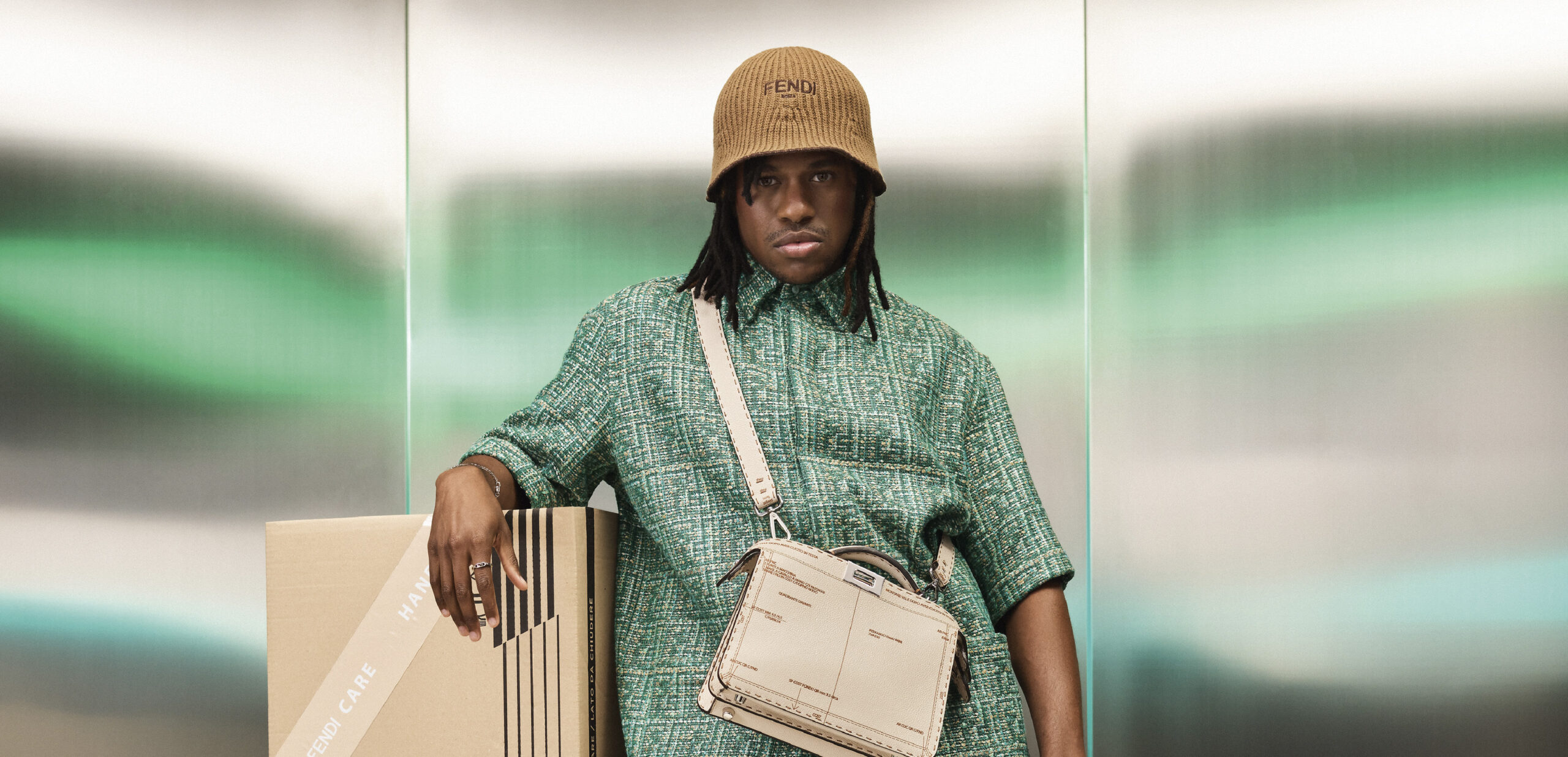 Jeremy Pope poses for Fendi Men's SS24, leaning on cardboard boxes, dressed in a patterned green jacket, khaki shorts, accessorized with a bucket hat, and carrying a white Fendi bag.