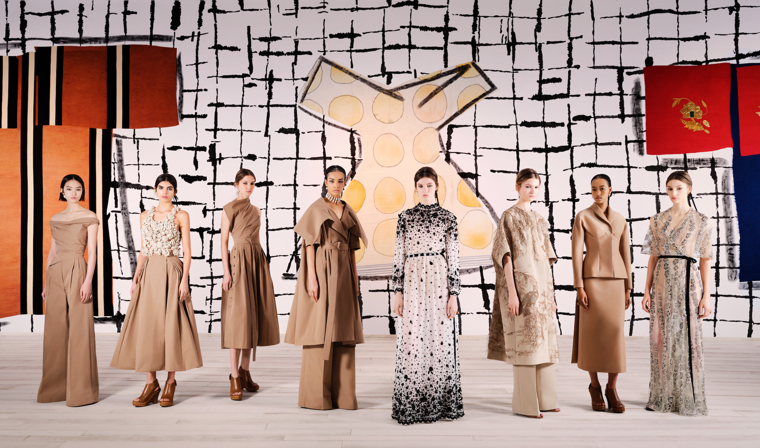 Models display Dior Spring 2024 Haute Couture collection, standing against a vibrant abstract wall, showcasing a range of elegant, neutral-toned outfits.