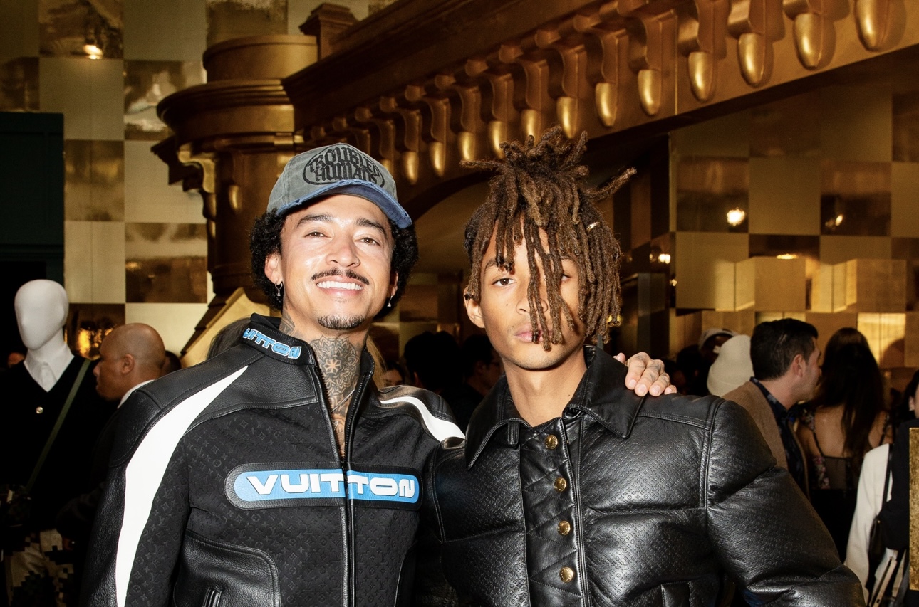 Nyjah Huston and Jaden Smith stand side by side, each showcasing their distinctive fashion sense. Huston is in a Louis Vuitton motorcross-inspired leather jacket, while Smith wears a textured black leather jacket with classic button details. Their shared smiles and effortless style exude a sense of camaraderie and youthful exuberance against the opulent backdrop of a well-appointed venue.