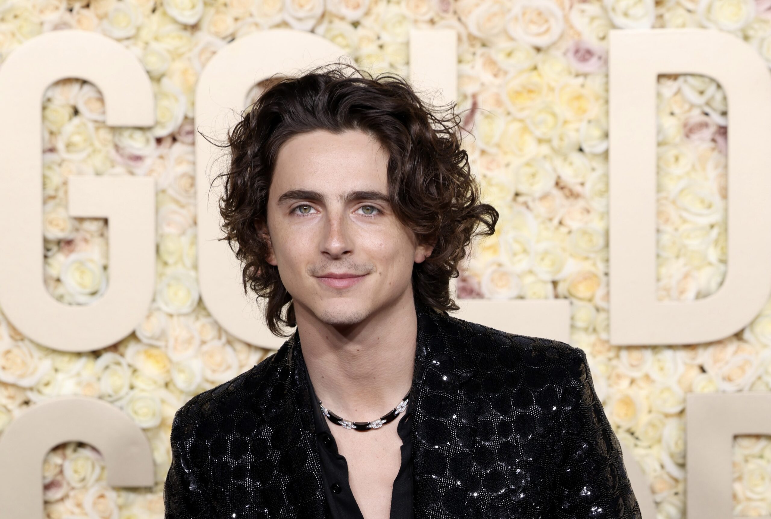 In a close-up at the Golden Globe Awards, Timothée Chalamet exudes elegance with a detailed view of his black sequined blazer, its pattern intricate and catching the light. His open-collar black shirt frames a statement silver necklace, and his poised expression reflects his status as a fashion-forward figure in the industry.