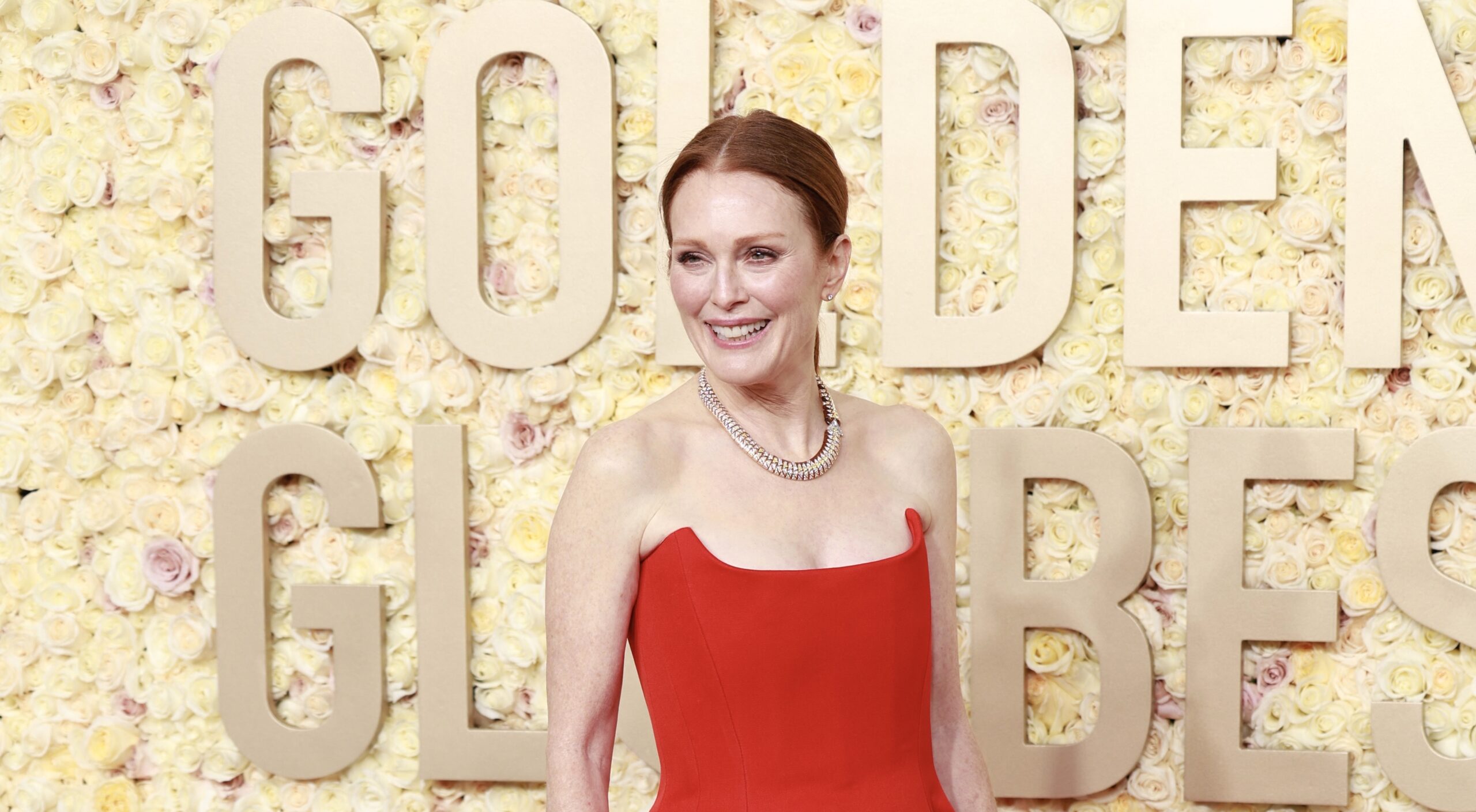 Julianne Moore stands gracefully on the red carpet at the 81st Golden Globe Awards, wearing a striking vermilion floor-length gown. The dress features a classic fit-and-flare silhouette with a strapless bodice, adding a timeless elegance to the event. Her necklace, a string of pearls, complements her attire, while her poised demeanor and light smile capture the essence of the evening's celebratory atmosphere. The background is adorned with a floral arrangement spelling 'GOLDEN GLOBES', enhancing the luxurious ambiance of The Beverly Hilton hotel in Beverly Hills, California.