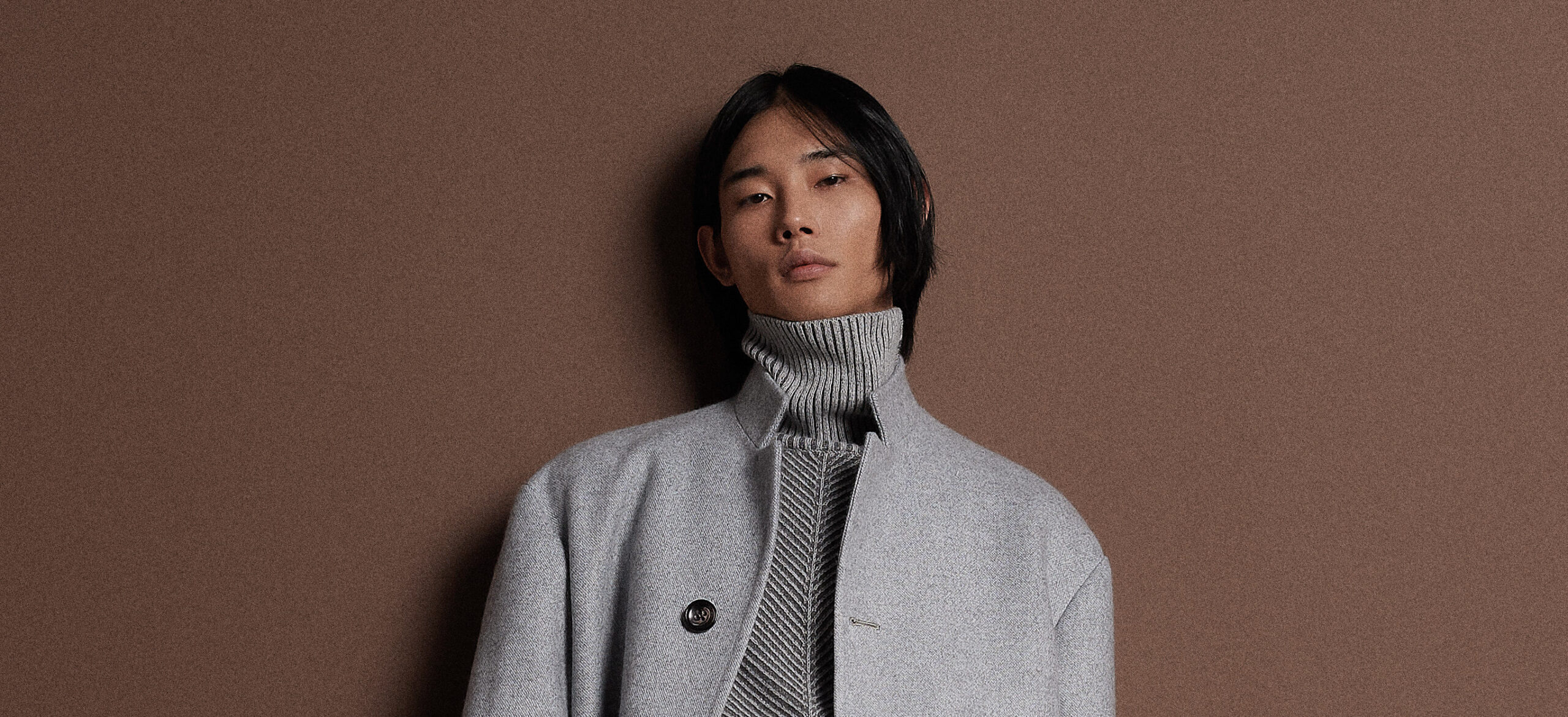 A model from KITON's AW24-25 collection stands against a warm earthy backdrop, sporting a grey turtleneck and blazer that epitomize the label's luxurious approach to classic men's fashion.