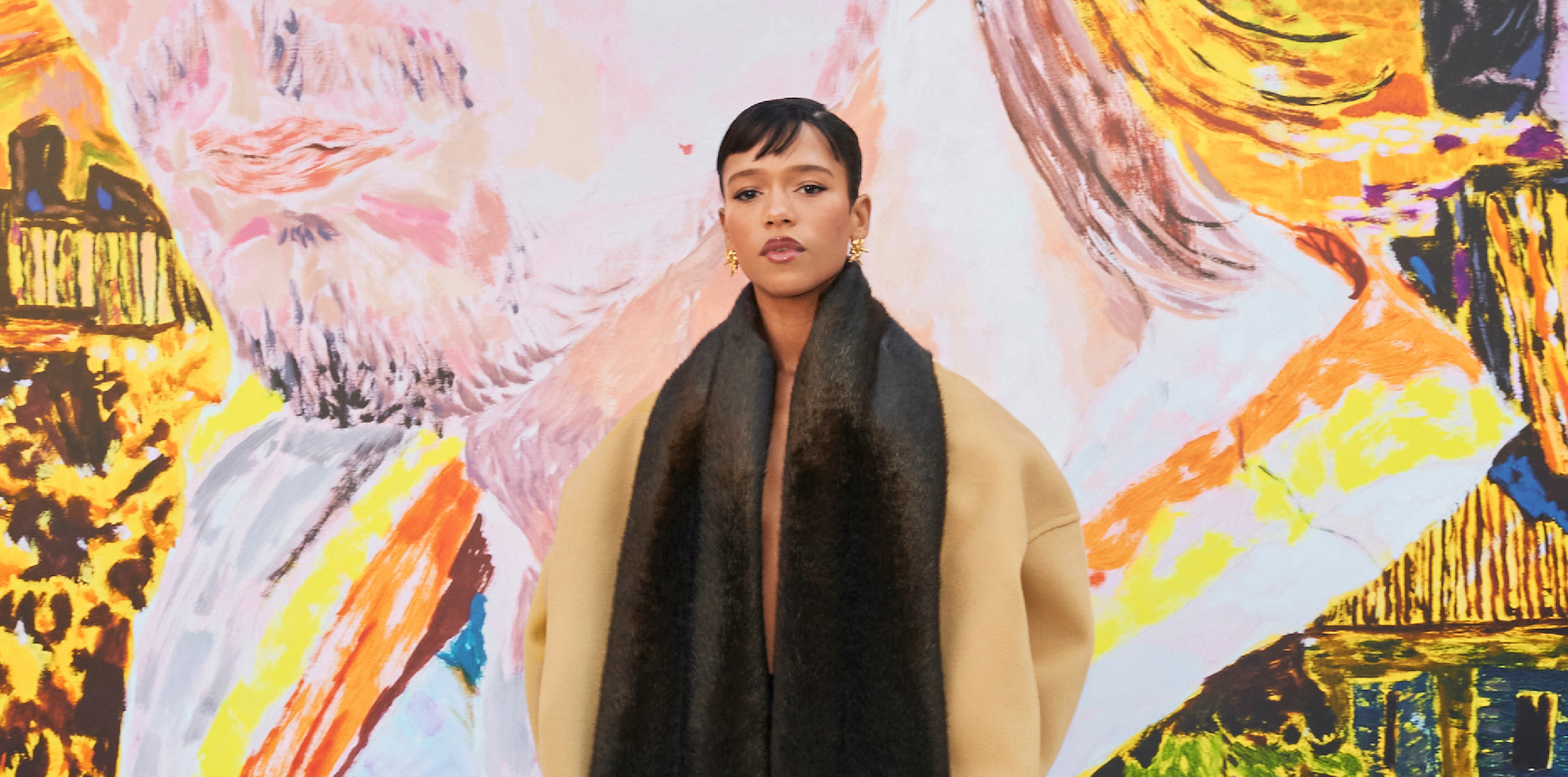 Actress Taylor Russell at LOEWE FW24 Men's Show, enveloped in a luxurious camel coat with a striking black scarf, against an abstract art backdrop.