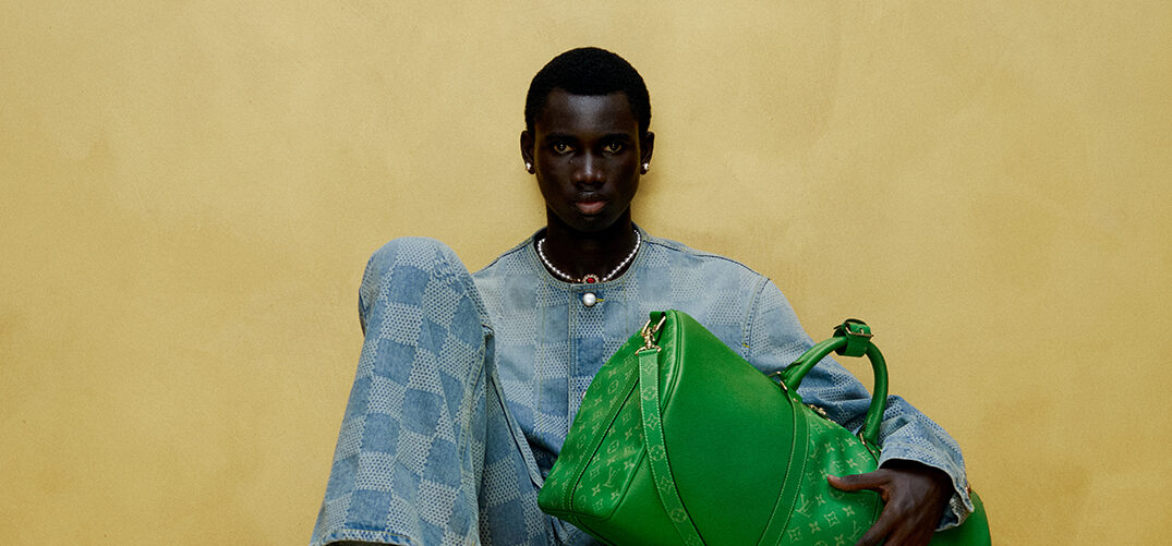 A model sits atop a classic Louis Vuitton trunk, clutching a vibrant green TAIGARAMA bag, wearing a denim-on-denim outfit with a checkerboard pattern, against a mustard yellow backdrop.