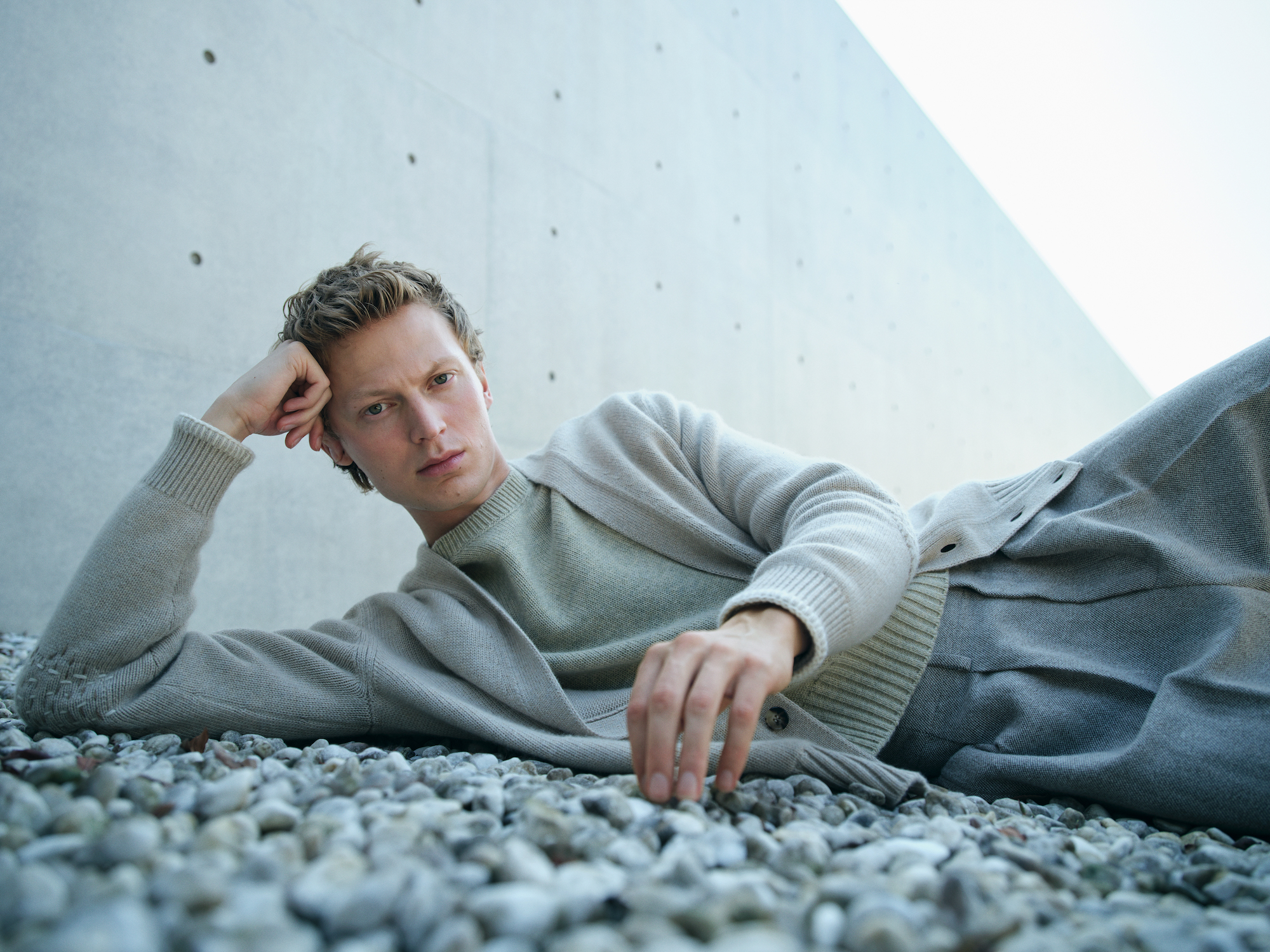 A male model lies on a pebble ground, leaning against a concrete wall, dressed in Loro Piana's comfortable knitwear and trousers, exuding a peaceful and stylish demeanor.