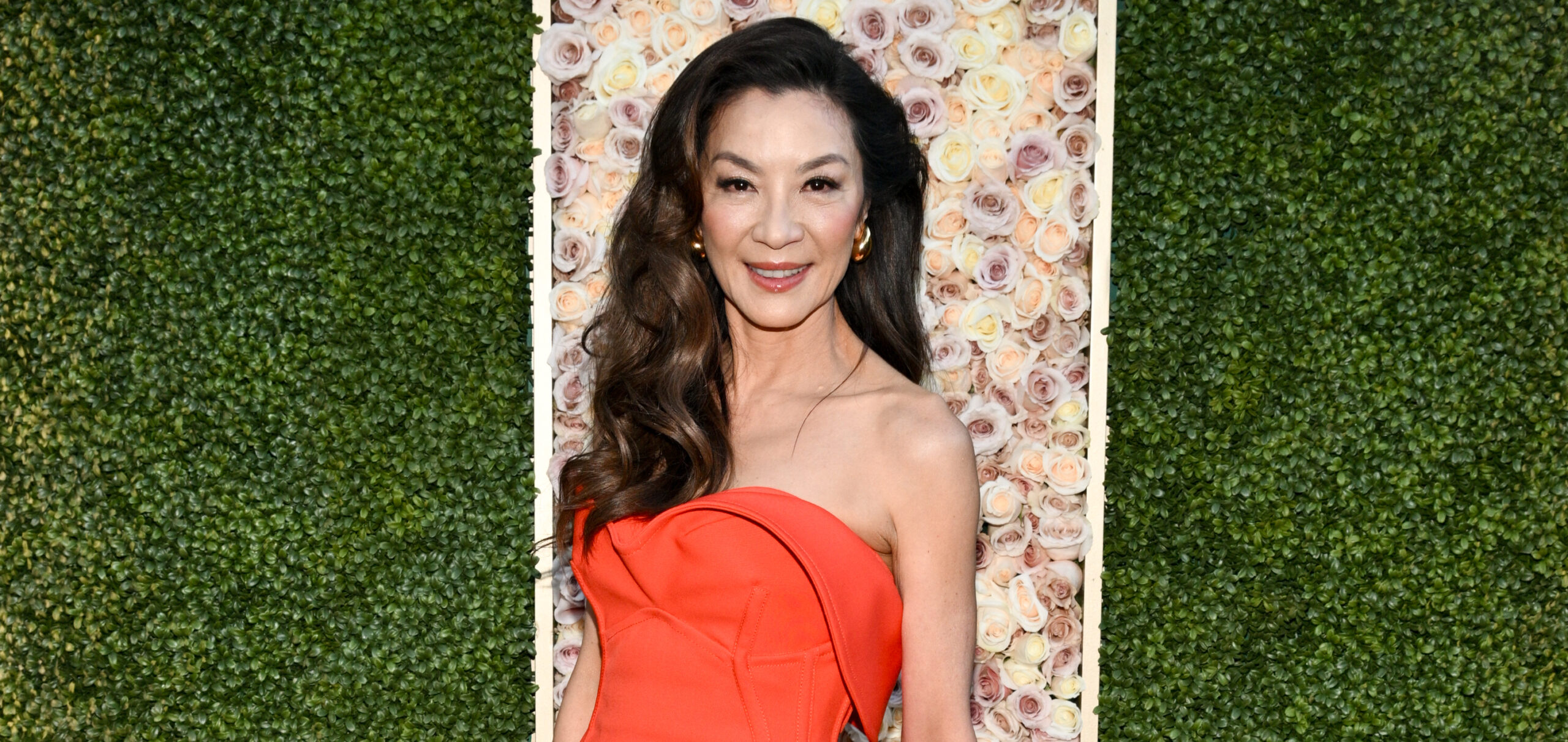 Michelle Yeoh graces the Golden Globe Awards in a stunning red strapless gown that speaks to her elegant poise. The gown's sleek silhouette gently traces her form, finishing with a subtle flair near the hem. Her ensemble is accessorized with a multi-colored, structured clutch and silver pointed heels, adding a playful yet sophisticated touch to her look. Her hair, swept back into a graceful style, showcases her warm smile and sparkling earrings, completing a picture of timeless beauty against the floral and verdant backdrop.