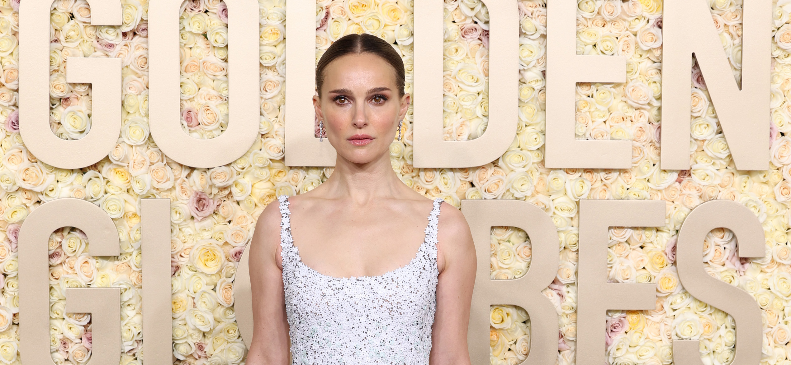 Natalie Portman stands at the 81st Annual Golden Globe Awards, embodying the essence of spring in a gown adorned with delicate, multicolored floral embellishments. The dress features a fitted bodice that transitions into a full, floor-grazing skirt, creating an ethereal silhouette. Her poised, straight posture and serene expression lend an air of refined sophistication, while the gown itself sparkles subtly, mirroring the floral backdrop of the event.