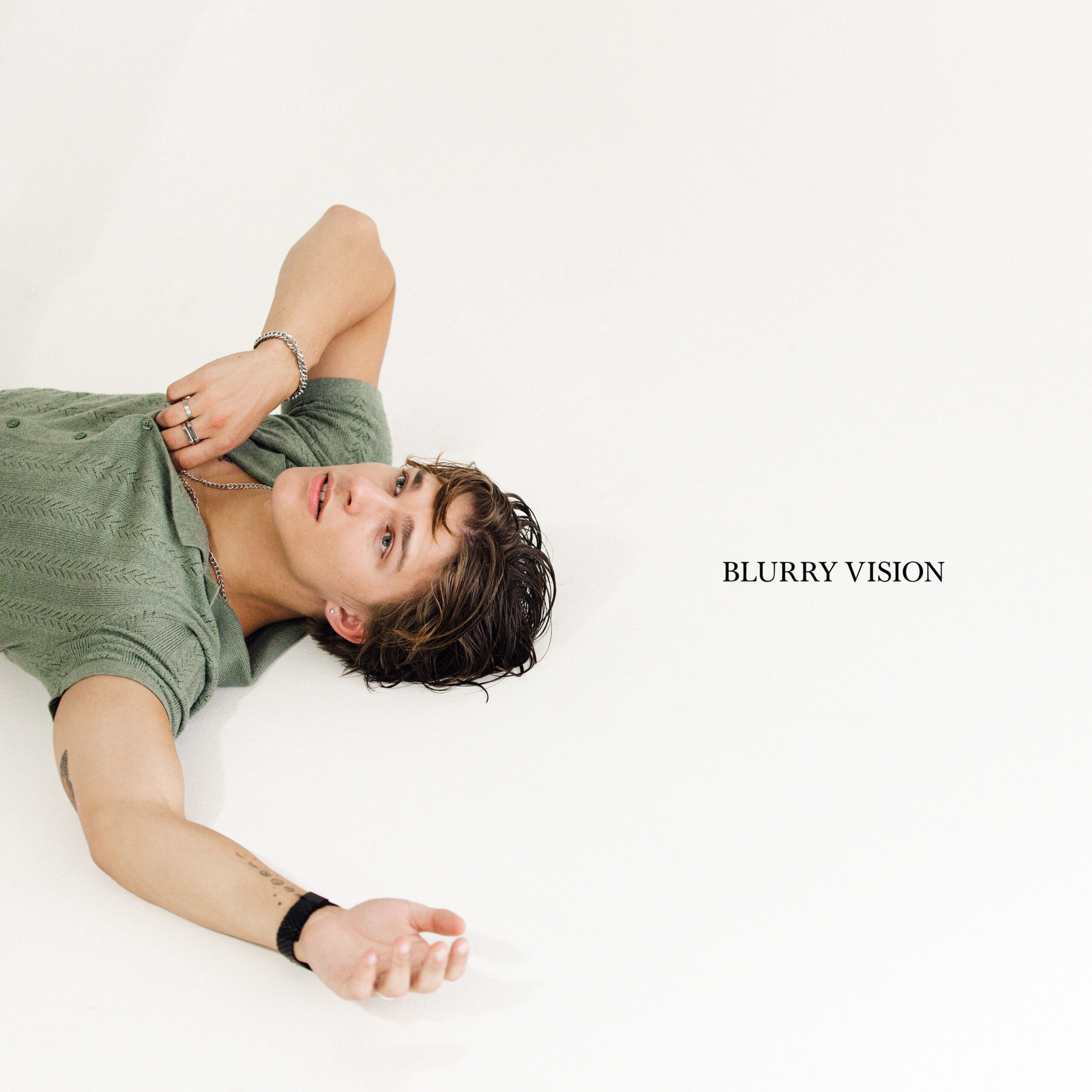 Alex Sampson lies on his back, gazing upward with a contemplative expression, wearing a sage green knit shirt. His wet hair is tousled, and he's accessorized with subtle jewelry. The phrase 'BLURRY VISION' is inscribed to the side, suggesting a deeper, introspective context.