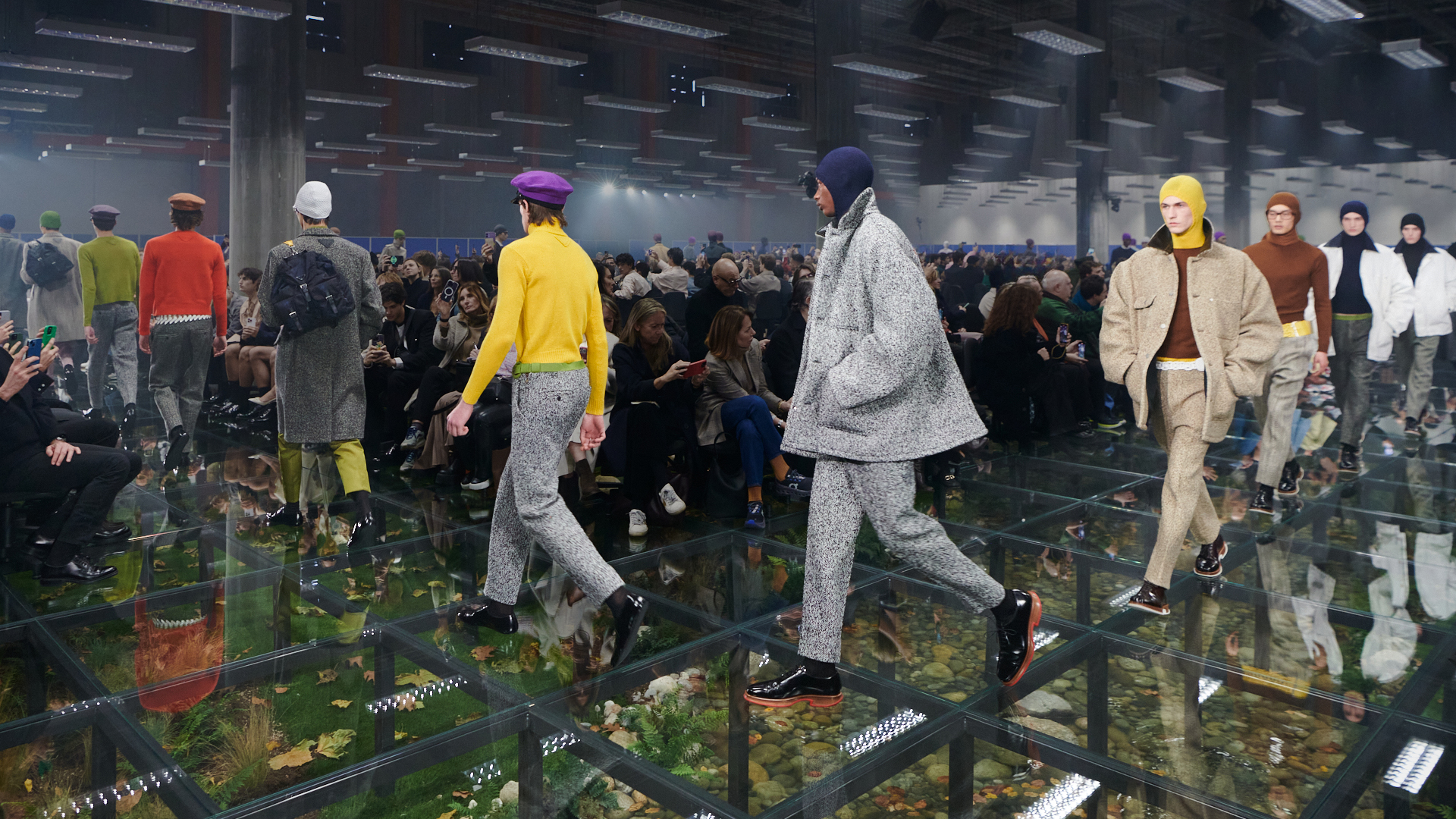 Models in the Prada Men's FW24 collection walking on a transparent glass runway over an artificial autumnal forest floor, with audience members on either side capturing the event. The fashion showcases a mix of vibrant and muted tones with varied textures, highlighted by distinctive Prada headwear.