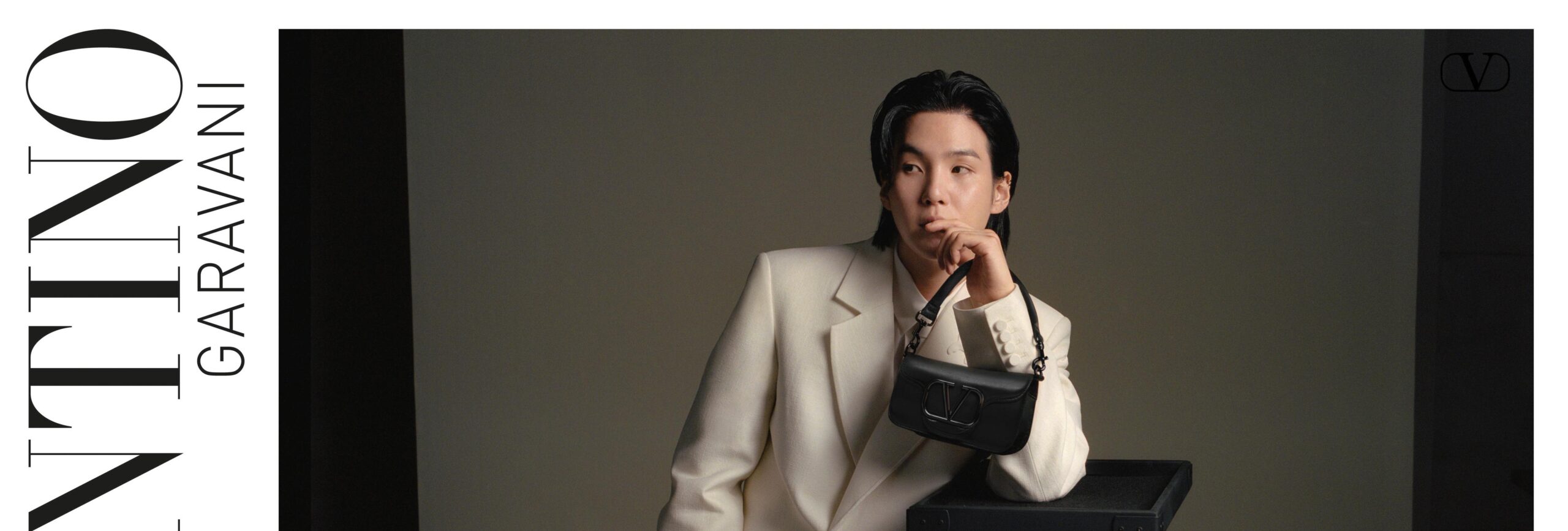 Suga of BTS poses with poise, leaning on a stand, adorned in an elegant beige suit and holding a small black handbag.