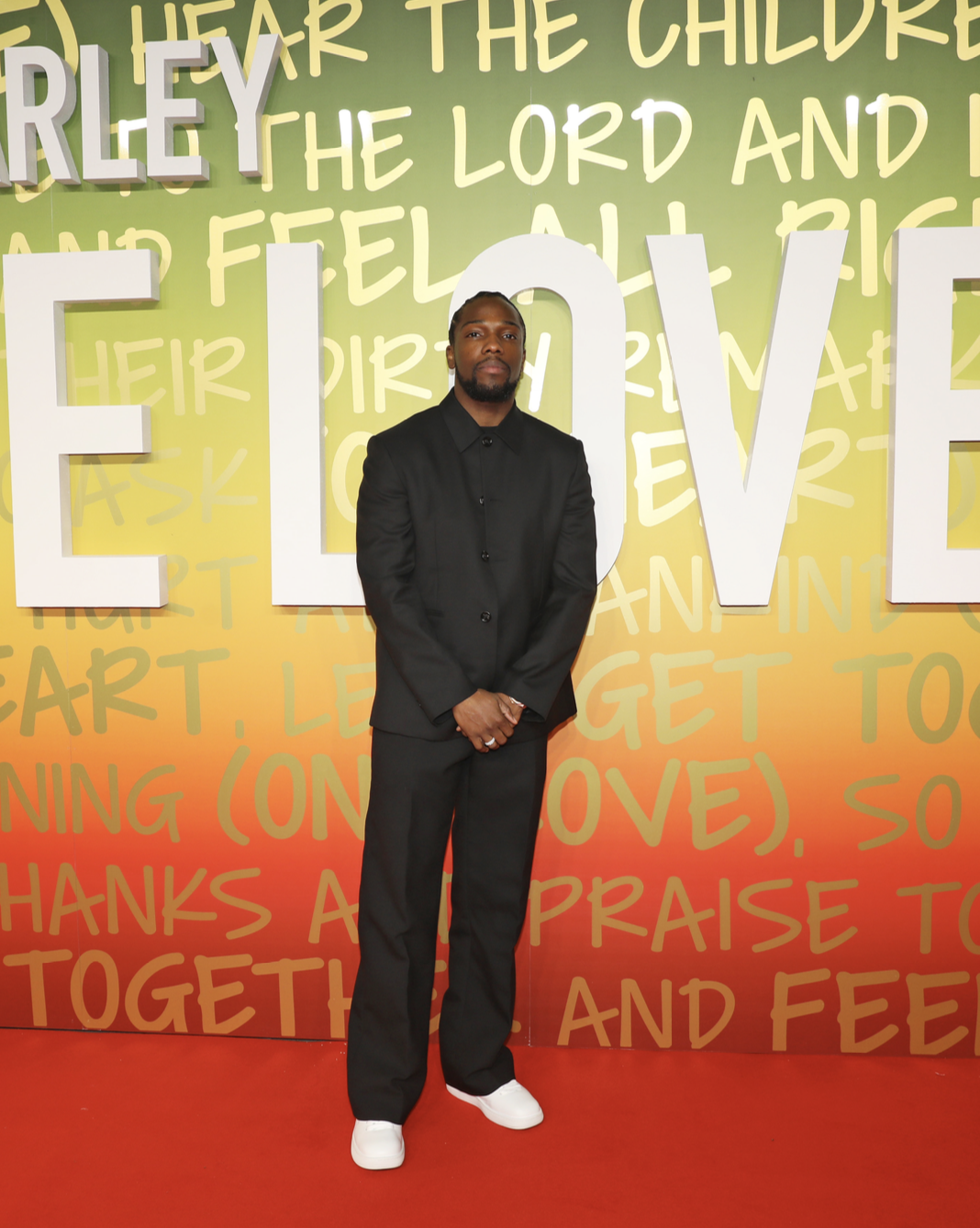 Tosin Cole stands confidently at the "One Love" premiere in London, wearing a bespoke Burberry black wool suit with white leather Box sneakers, against a vibrant backdrop with bold lettering.