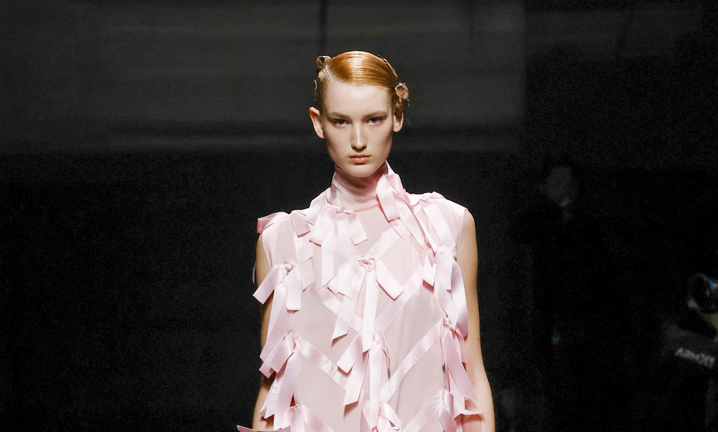 A model showcases a Prada Fall/Winter 2024 collection piece on the runway, featuring a pale pink sleeveless dress adorned with ribbon-like details, complemented by a high neck and structured silhouette.