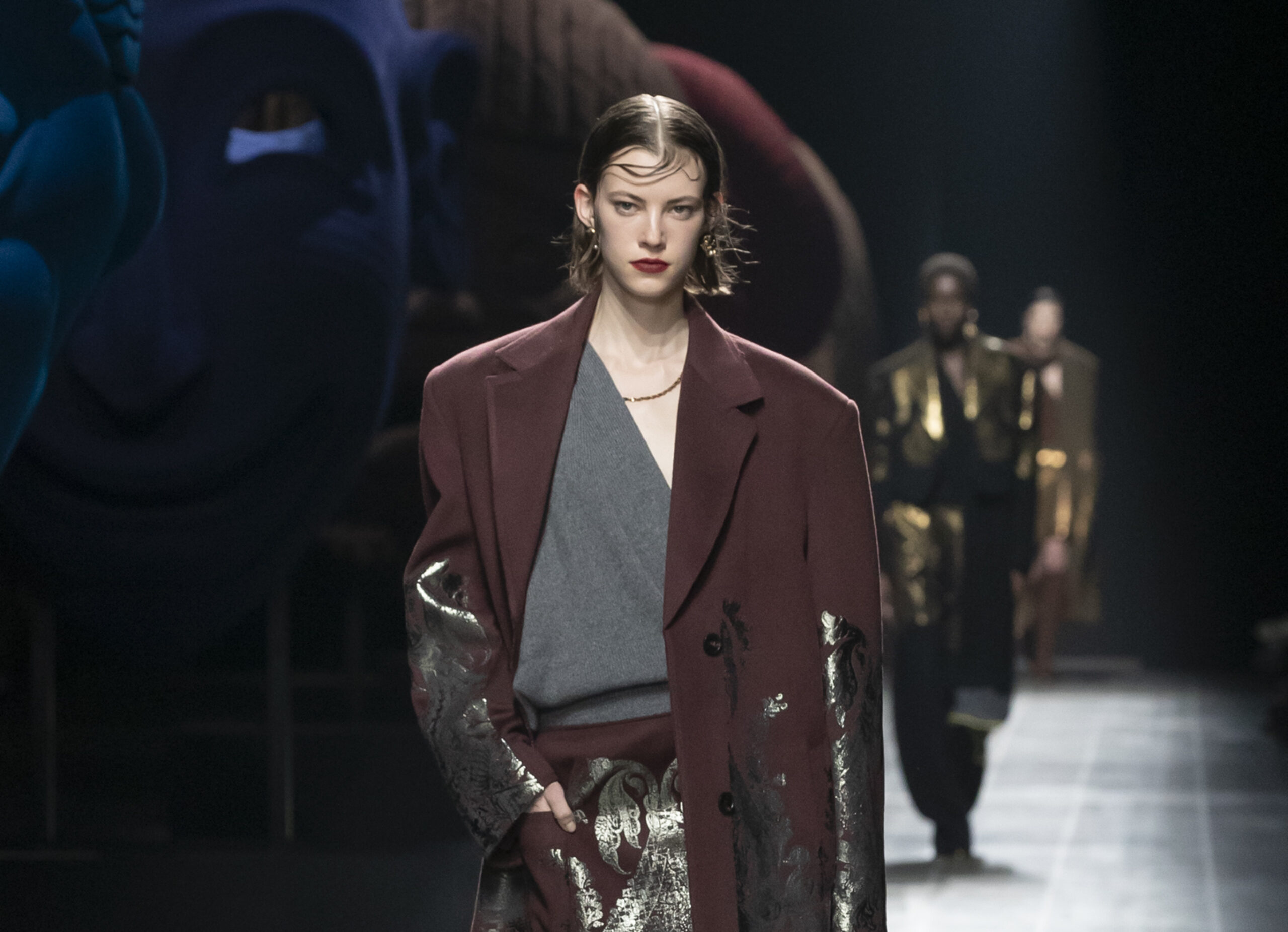 A model walks the runway in a layered outfit from Etro's Fall/Winter 2024/2025 collection, featuring a maroon coat with silver accents.