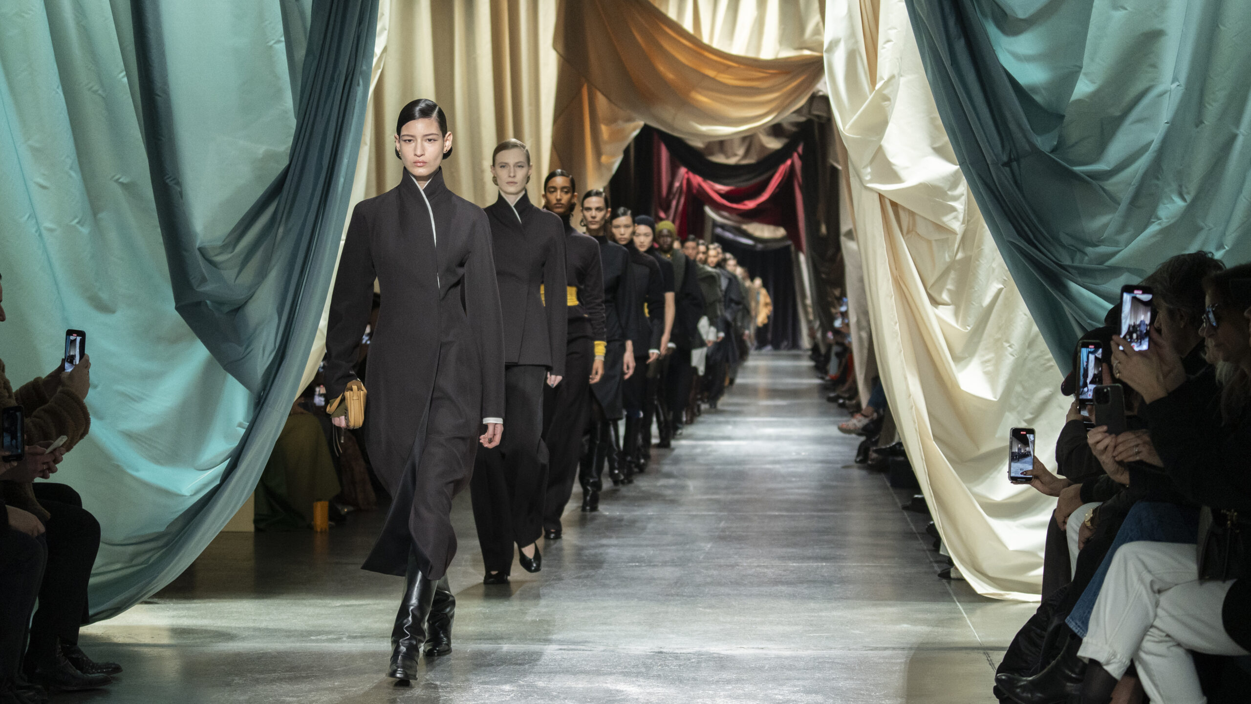 A procession of models striding down the runway in a fashion show, clad in Fendi's Autumn/Winter 2024-25 collection. The opulent setting features draped fabrics in pastel and neutral tones along the catwalk, with an audience capturing the moment on their smartphones.