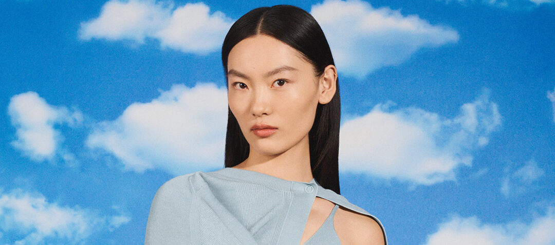 Close-up of a model against a sky backdrop, wearing a ribbed blue top with cut-out details and a brown glove, accessorized with a gold earring.