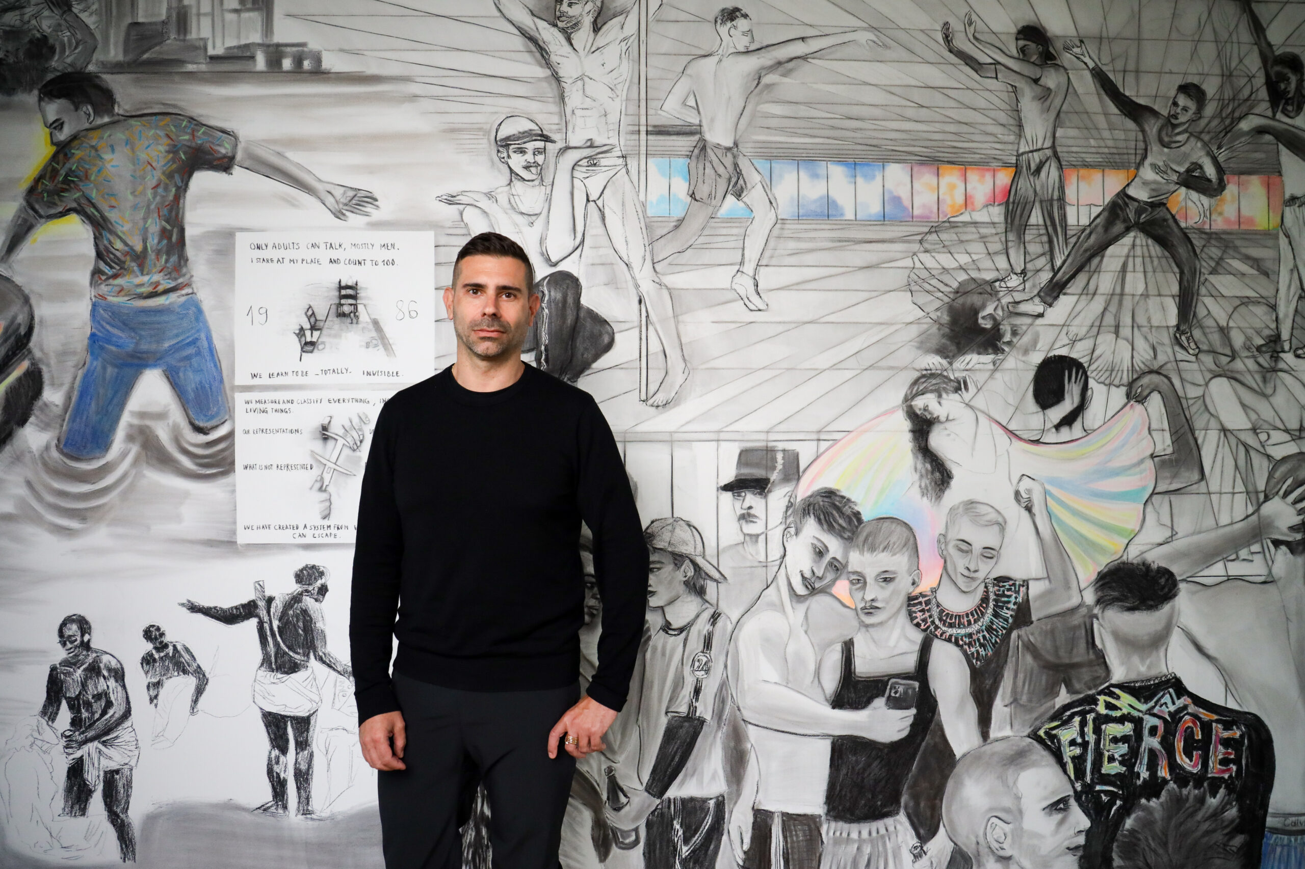Marc Bauer stands in front of a detailed mural, dressed in a black long-sleeve shirt and black pants, offering a stark contrast to the lively and intricate grayscale artwork behind him that depicts a variety of human figures and text elements in a dynamic composition.