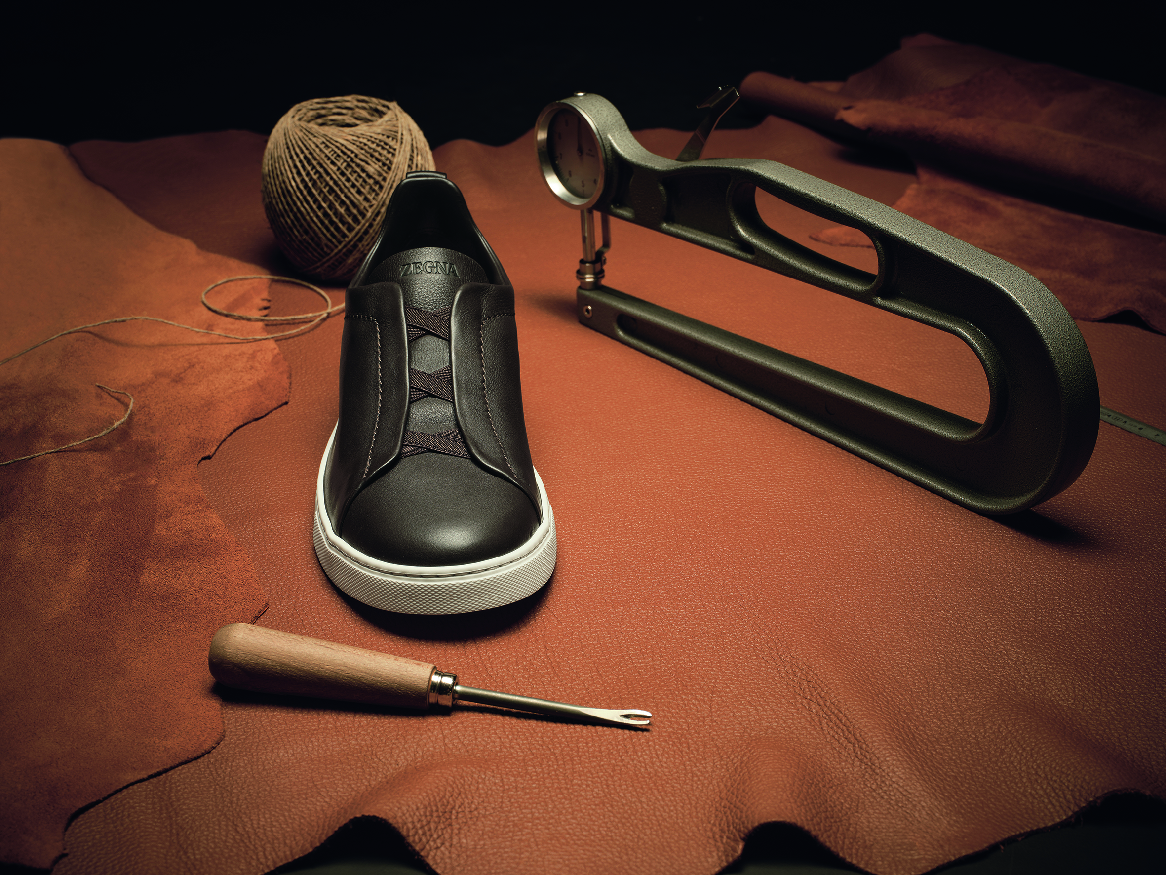 A completed black Zegna Triple Stitch Secondskin shoe is placed on a large piece of tawny leather, surrounded by a spool of thread, a shoemaker's awl, and a metal shoe stretcher in a dimly lit workshop setting.