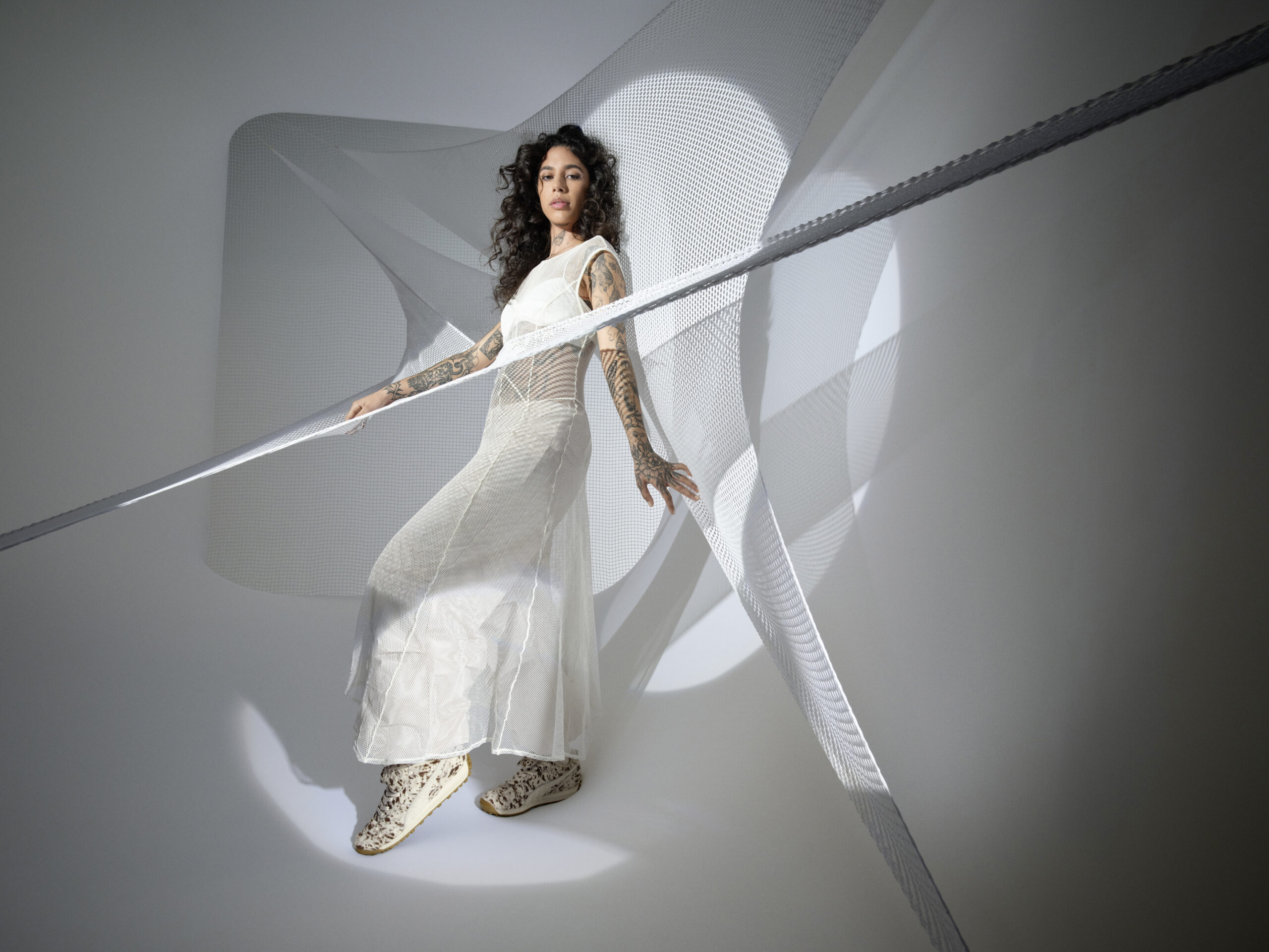 A model with elaborate tattoos on her arms poses in a flowing white dress and FENTY x PUMA Avanti Pony sneakers. The background is an abstract play of light and shadow with dynamic white ribbons crisscrossing around her, creating a sense of movement and modern elegance.