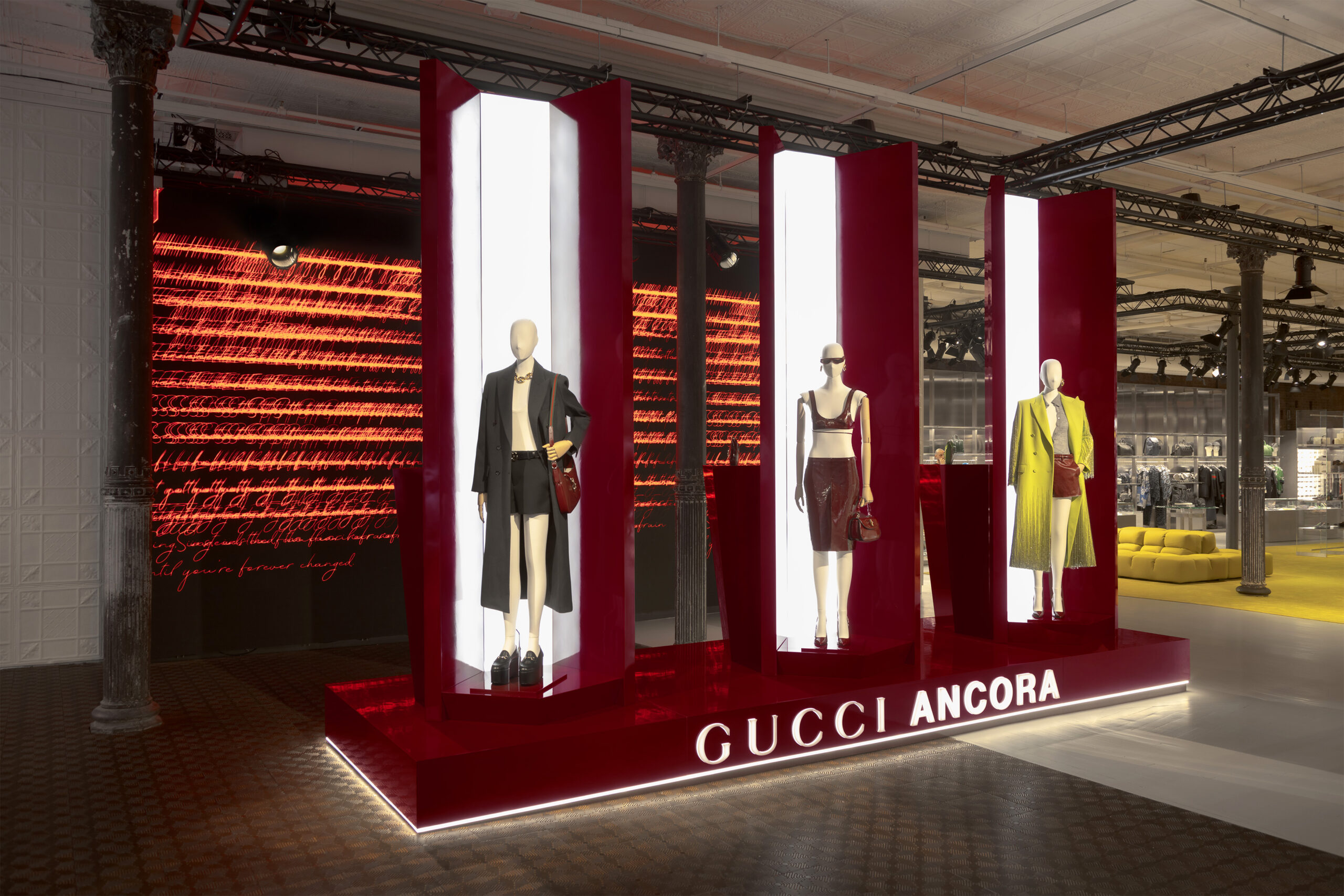 An elegant display inside the Gucci Wooster boutique featuring three mannequins showcasing the latest fashion designs against a backdrop of vibrant red hues and artistic lighting. The display is highlighted by the illuminated 'GUCCI ANCORA' sign at the base.