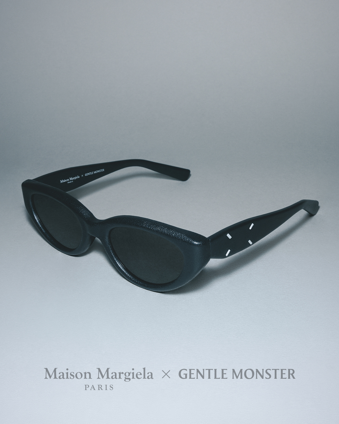 A pair of Maison Margiela x GENTLE MONSTER black sunglasses with distinctive leather-wrapped frames and four white stitches on the arm, resting on a grey background with their brand collaboration name displayed below.