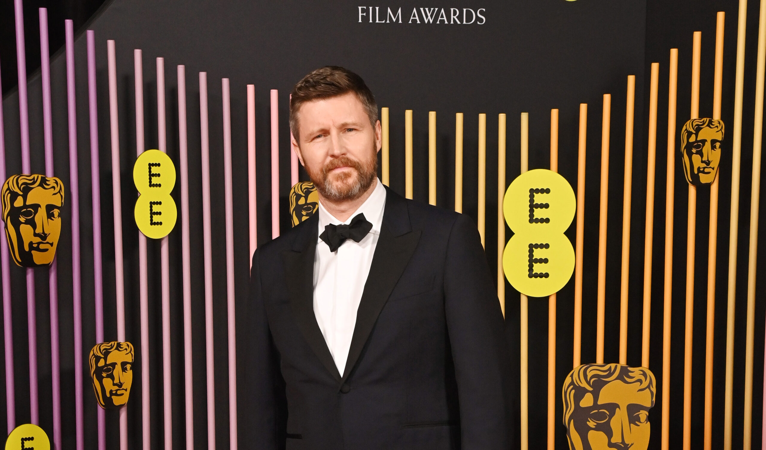 Andrew Haigh stands confidently at the 2024 EE BAFTA Film Awards, wearing a dark navy wool mohair tuxedo by dunhill, paired with a white cotton pique shirt and a black silk faille bow tie.