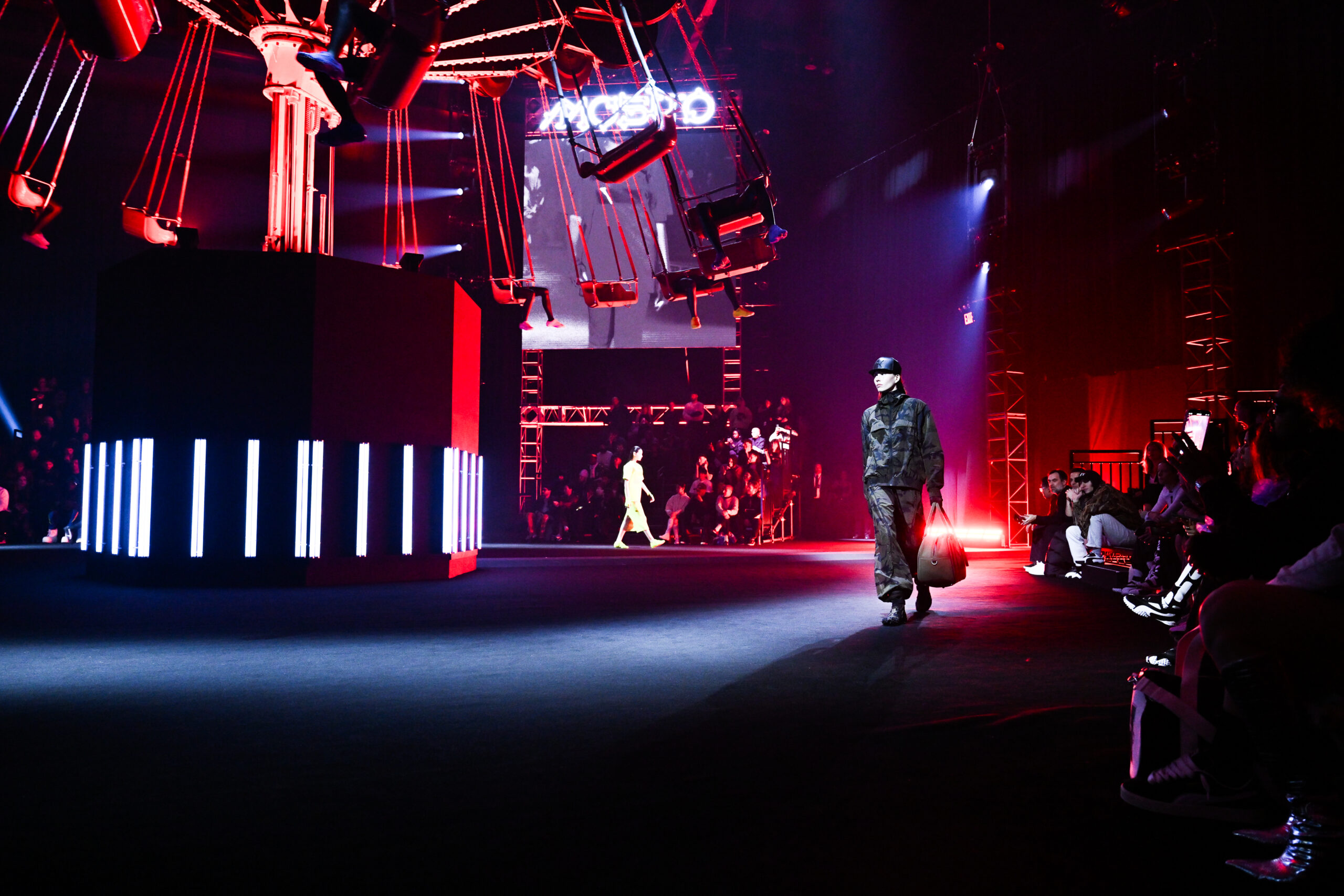 A dynamic fashion show scene with models walking the runway under vibrant red lights, featuring a setup titled "WELCOME TO THE AMAZING MOSTRO SHOW" presented by PUMA.