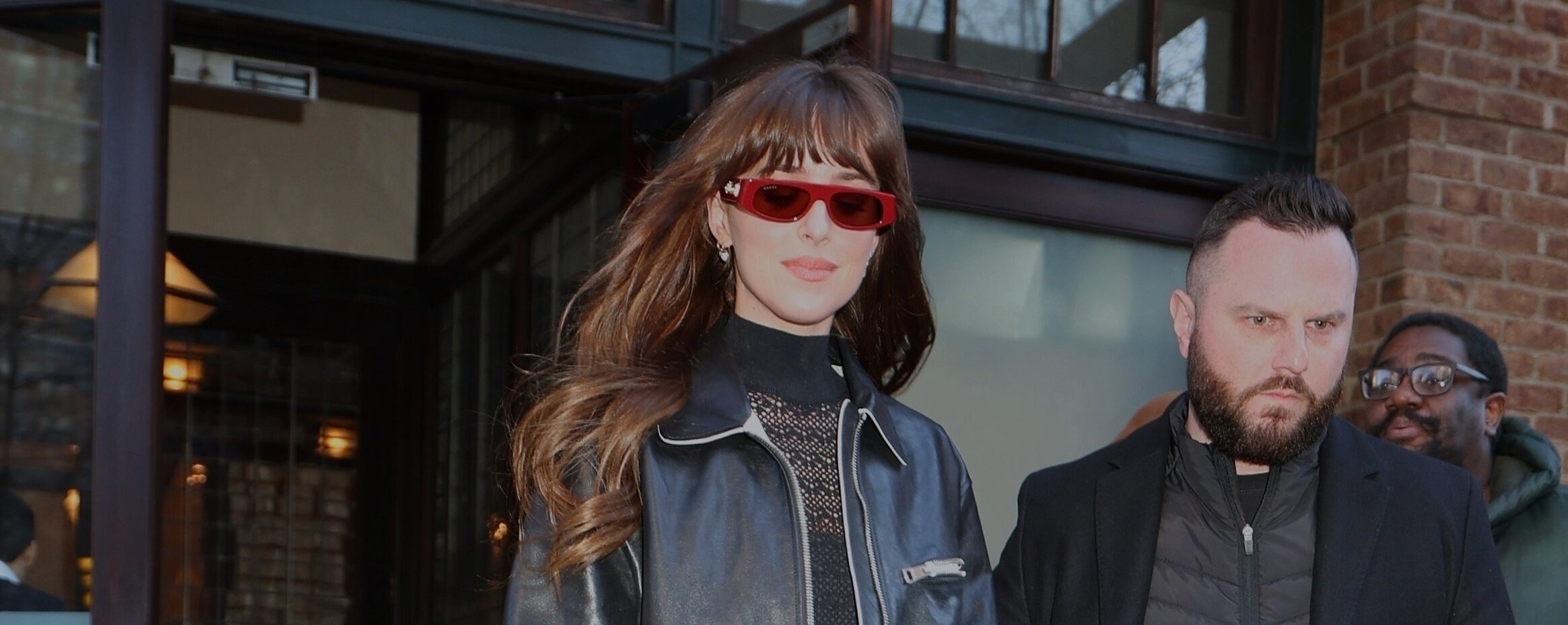 Dakota Johnson steps out onto a city street dressed in a black leather jacket over a black turtleneck and a shimmering long skirt. She accessorizes with red sunglasses, a matching red handbag, and black heeled boots, exuding a chic, modern look.