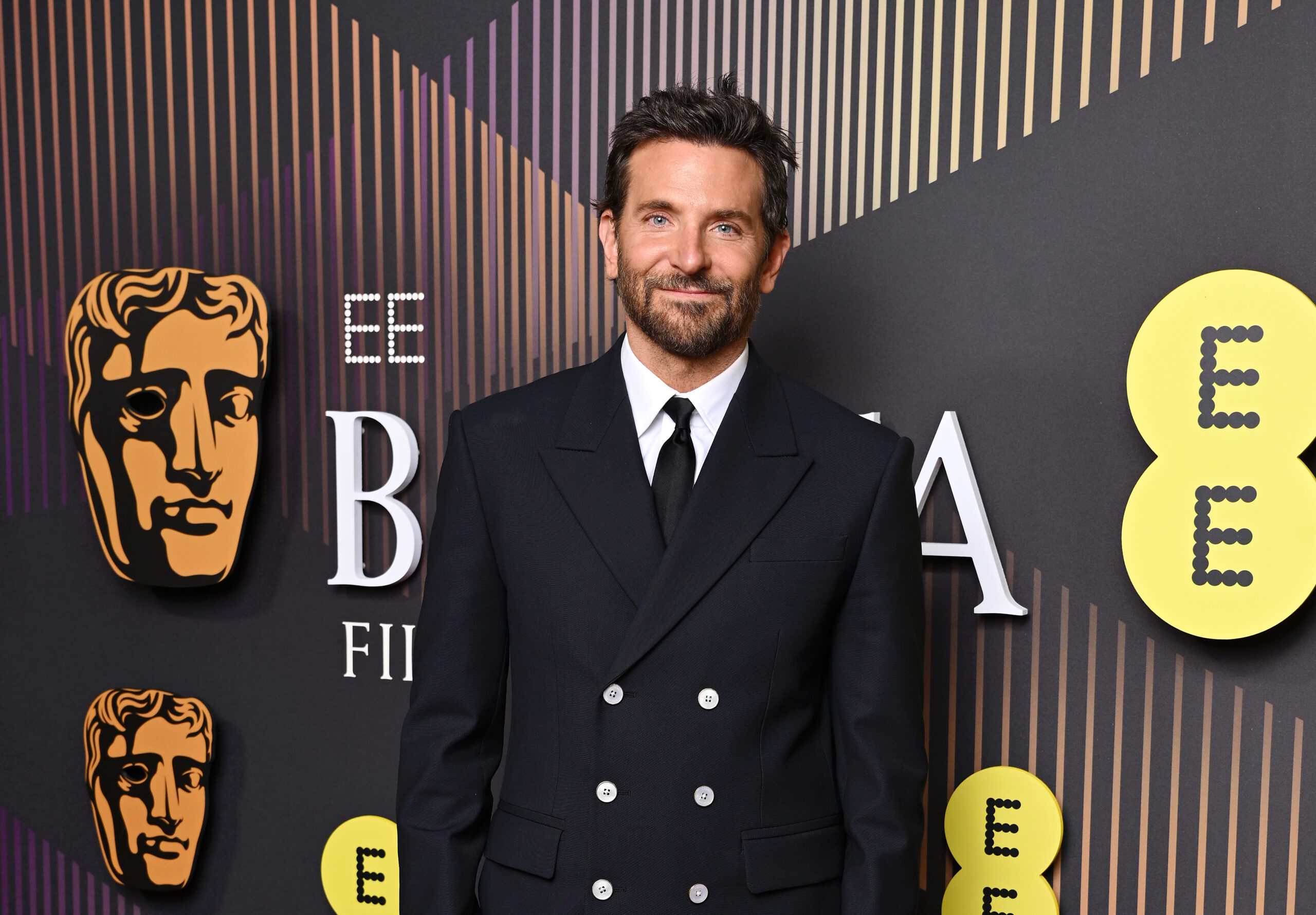 Bradley Cooper stands confidently at the 2024 BAFTA Film Awards, dressed in a custom Louis Vuitton ensemble featuring a double-breasted black coat, flared trousers, a white shirt, and a black tie, with the maison's luxury wristwatch on display.