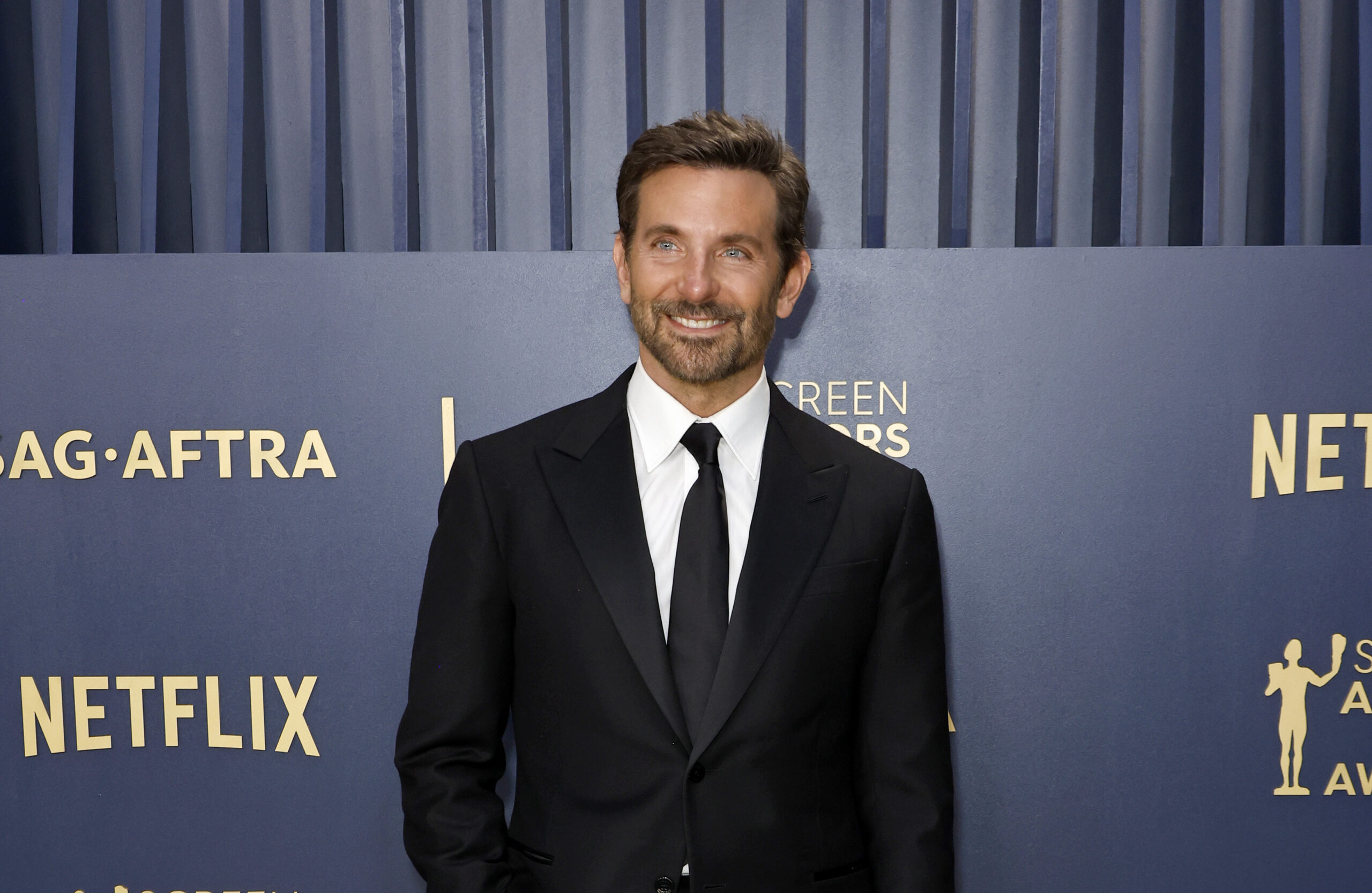 Bradley Cooper stands with poise on the red carpet at the 30th Annual Screen Actors Guild Awards, sporting a sharp black Louis Vuitton tuxedo paired with a classic white shirt and black tie. His attire is polished off with black derbies and a sophisticated steel Tambour watch with a navy dial, encapsulating a blend of classic and contemporary style.