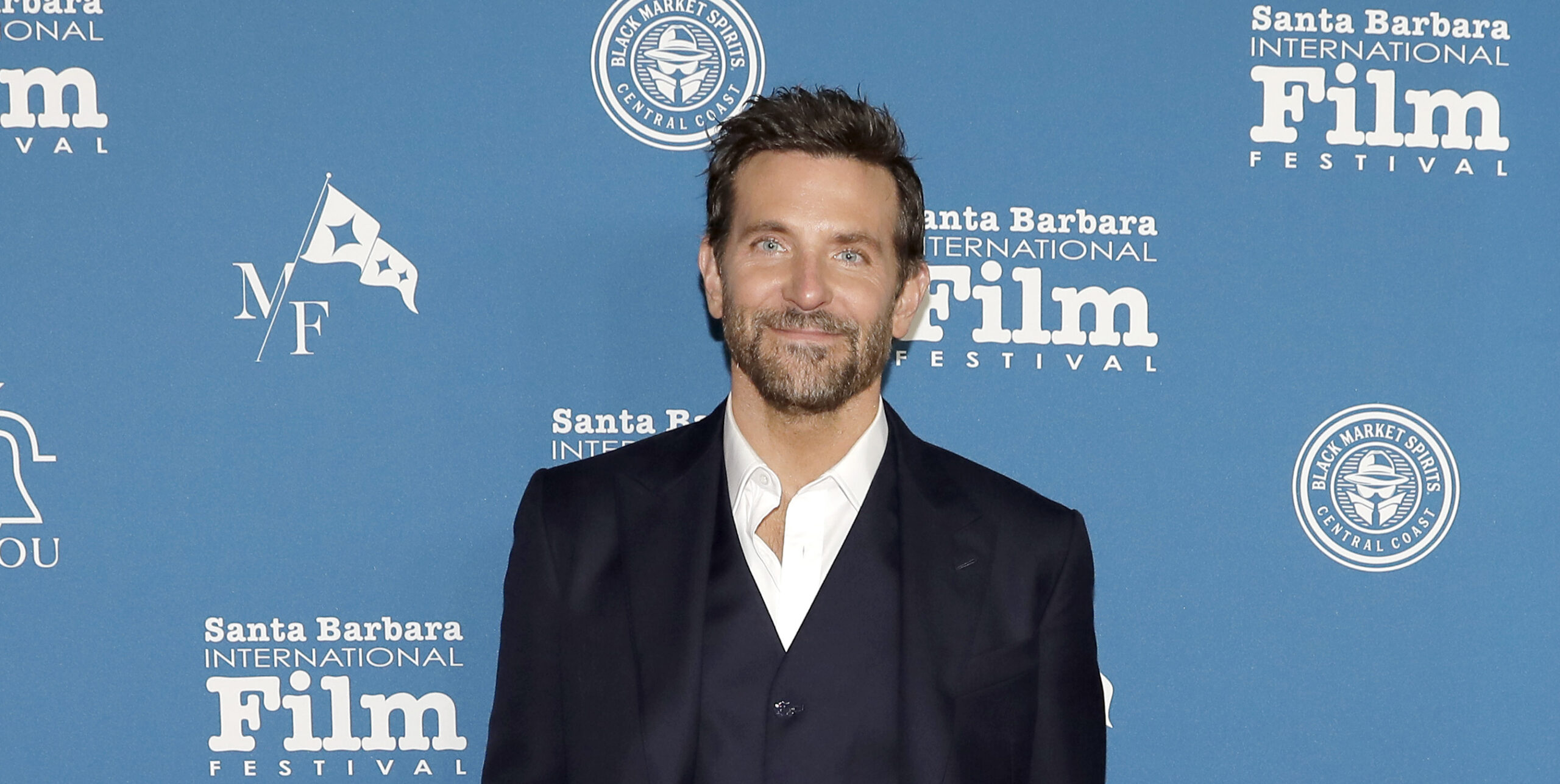 Bradley Cooper in a tailored Louis Vuitton navy suit stands on a red carpet against a backdrop with logos for the Santa Barbara International Film Festival.