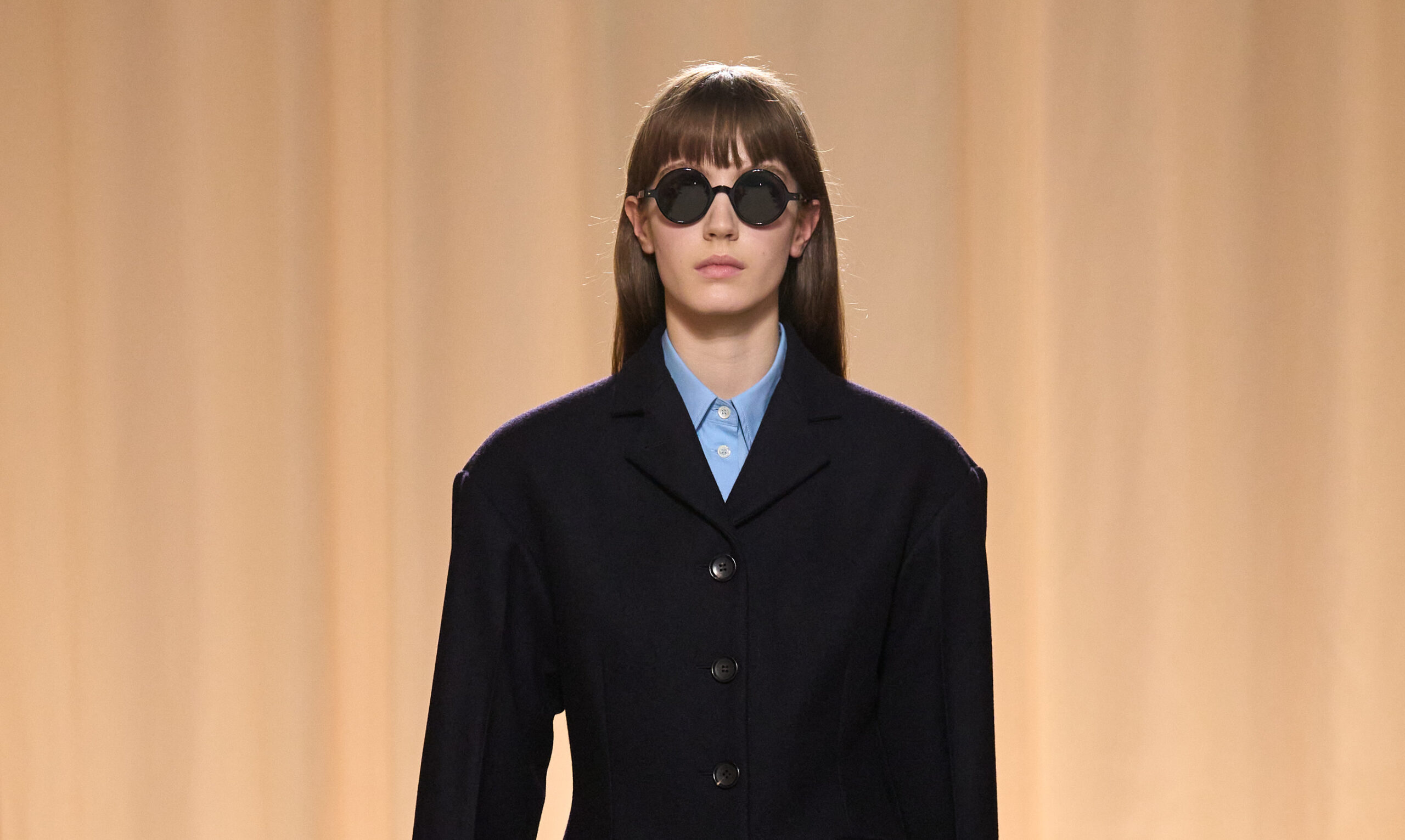 A close-up of a model in a Bally Fall/Winter 2024 fashion show, wearing a navy coat over a light blue shirt, accessorized with large round sunglasses.