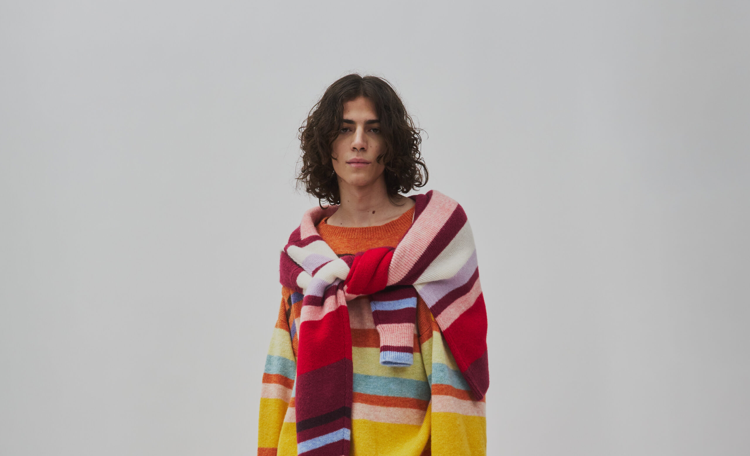 A model poses in an oversized, multicolored knit ensemble from Bulan's 'Garbage Collector' collection. The piece features a mix of warm and cool stripes, with hues of red, orange, yellow, blue, and white, creating a cozy yet striking visual appeal.